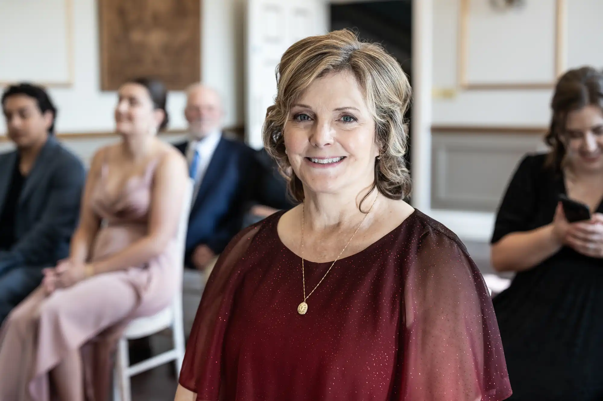 A woman in a burgundy dress smiles at the camera. Behind her, several people are seated, with one woman looking down at her phone.