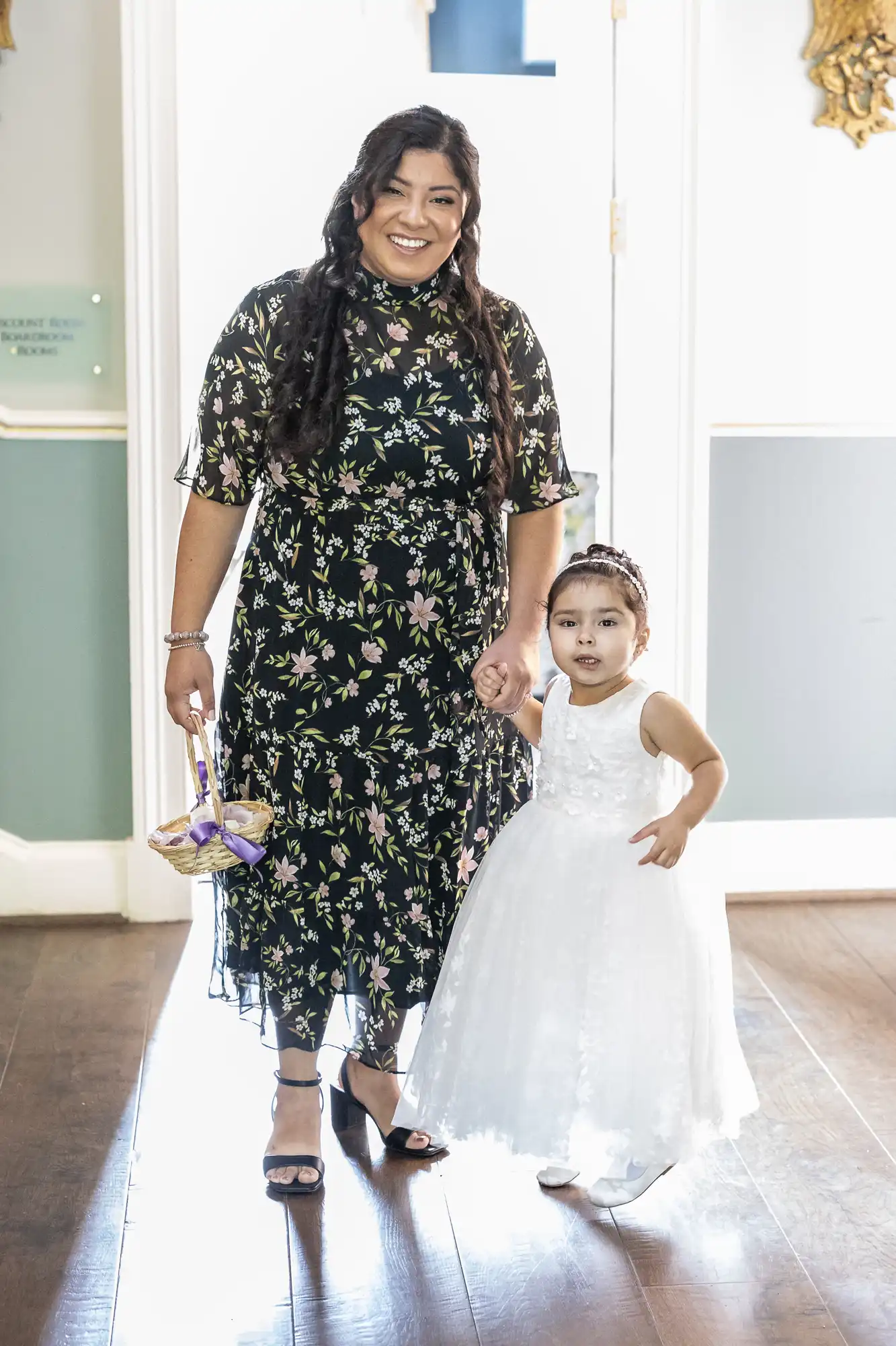 A woman in a floral dress smiles while holding the hand of a young girl in a white dress and headband inside a well-lit room. The girl holds a small basket.