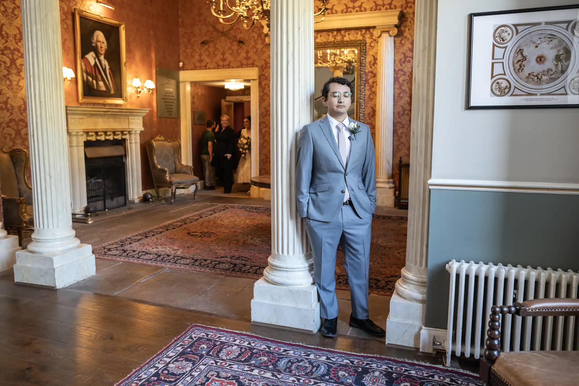 A man in a light gray suit and pink tie stands next to a column in a lavish room with ornate decor, including a portrait, chandelier, and patterned rugs. Other people are visible in the background.