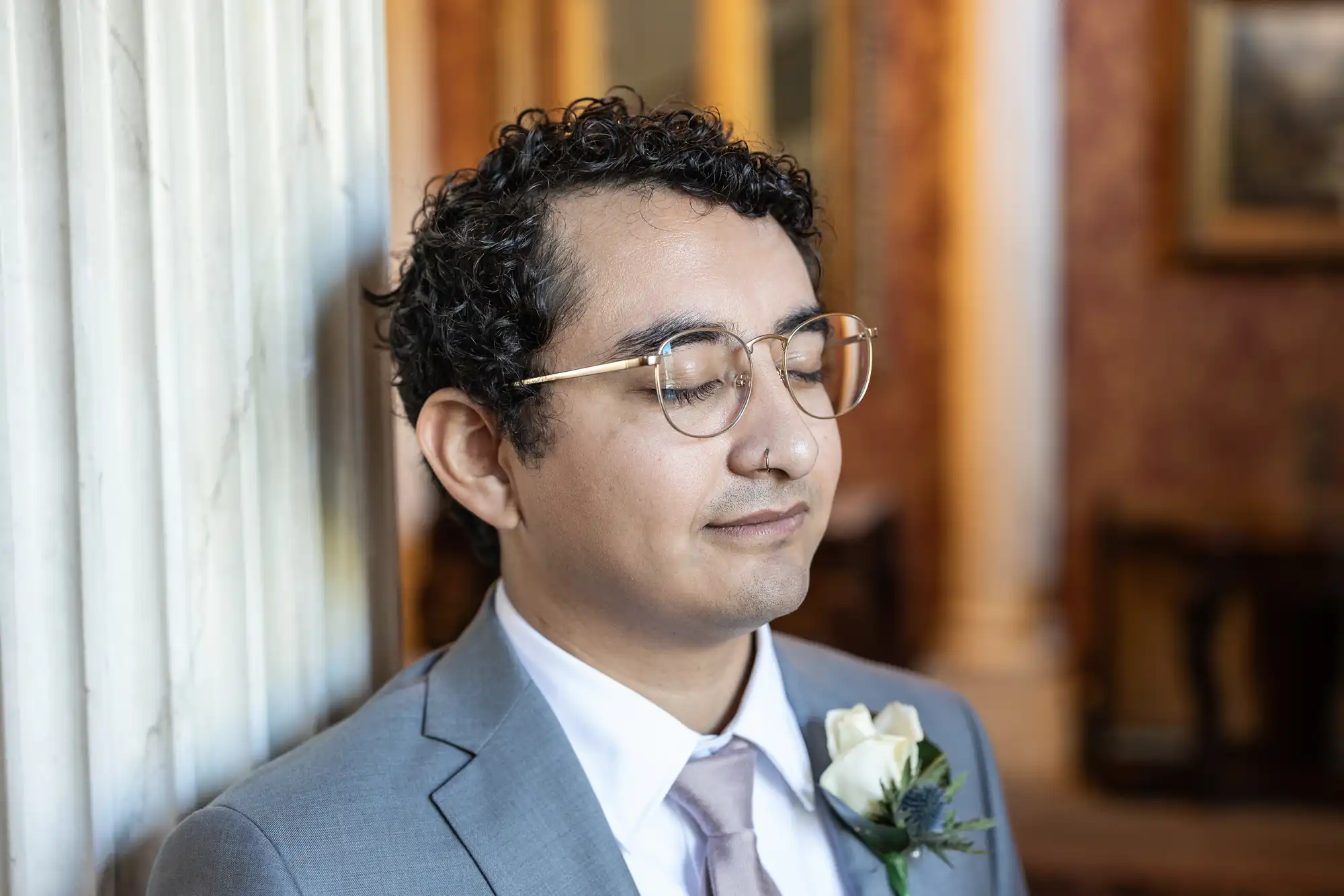 A man with curly hair, wearing glasses and a gray suit with a white rose boutonniere, stands with his eyes closed against a pillar in an indoor setting.