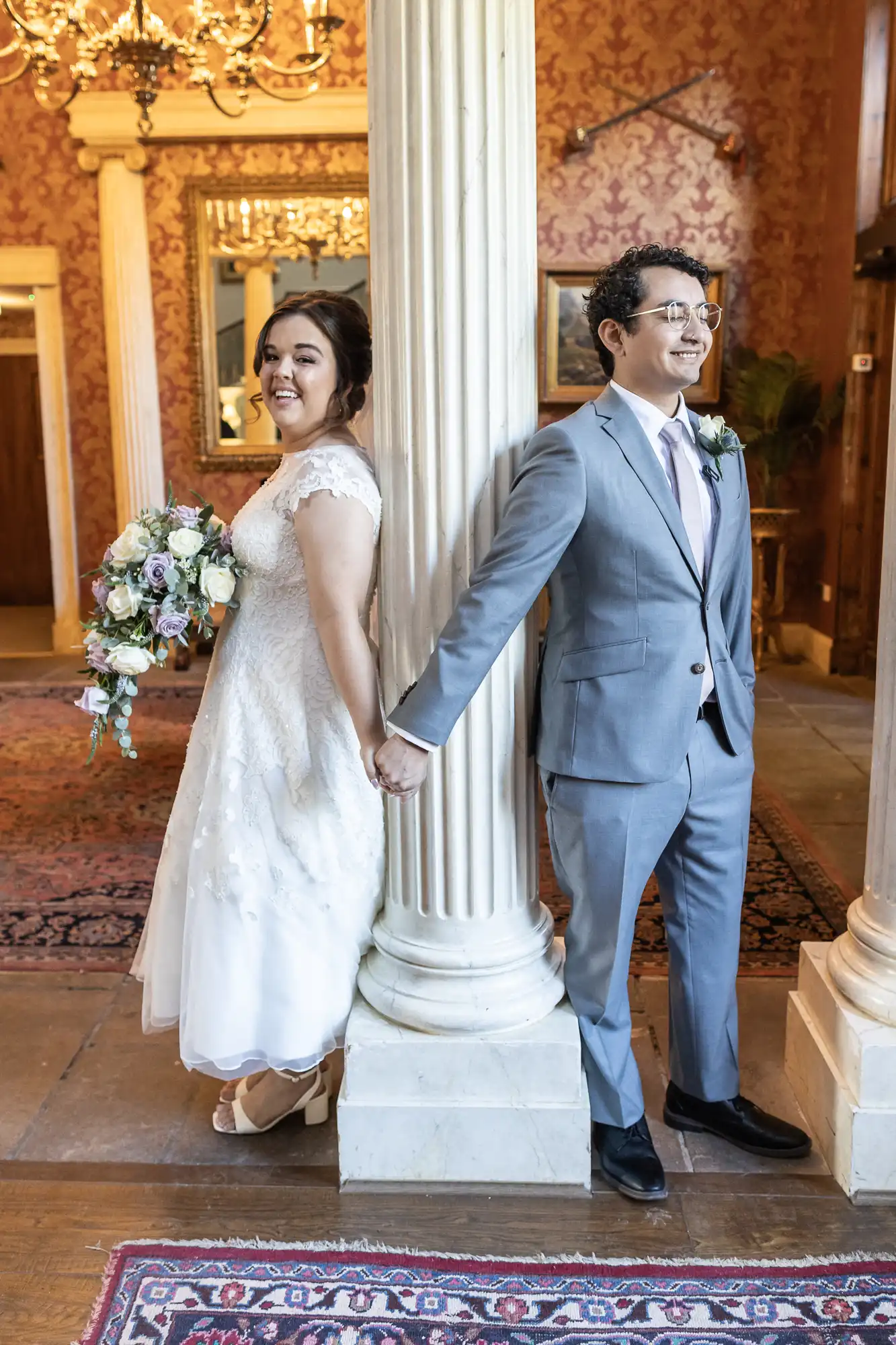 A bride and groom stand back-to-back, holding hands around a column. She wears a white lace dress and holds a bouquet; he wears a gray suit and a pink tie. They are indoors, in an ornate setting.