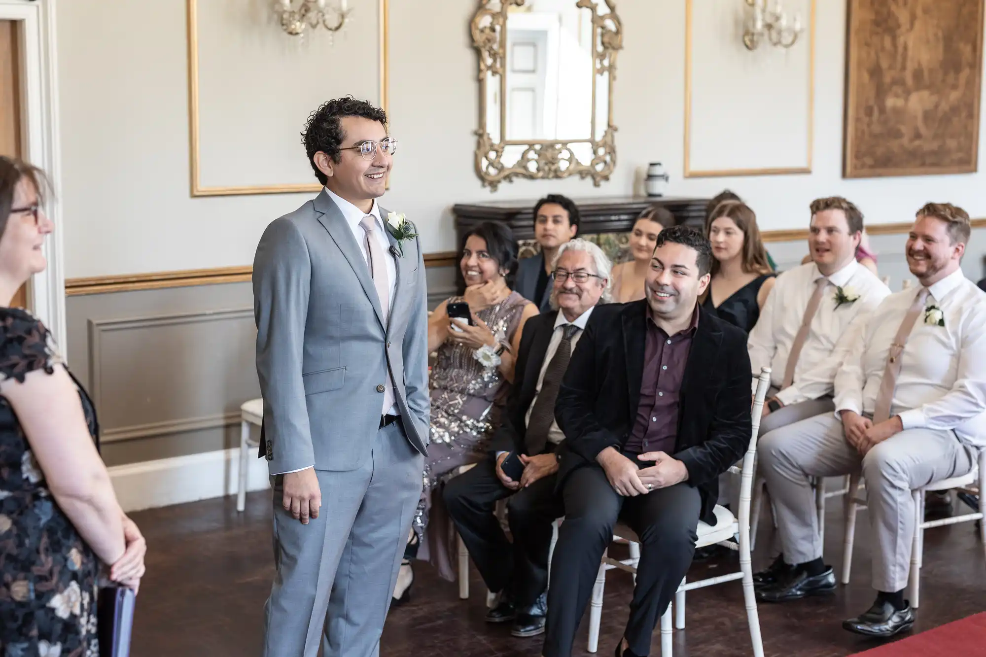 A man in a gray suit stands smiling at the front of a room while guests seated in rows look on, some smiling, during a ceremony.