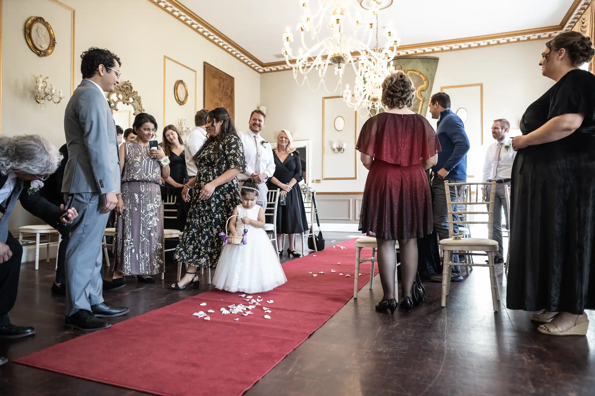 A young girl in a white dress walks down a red carpet aisle strewn with petals. People, some in formal attire, stand on either side in a decorated room with chandeliers and paintings.