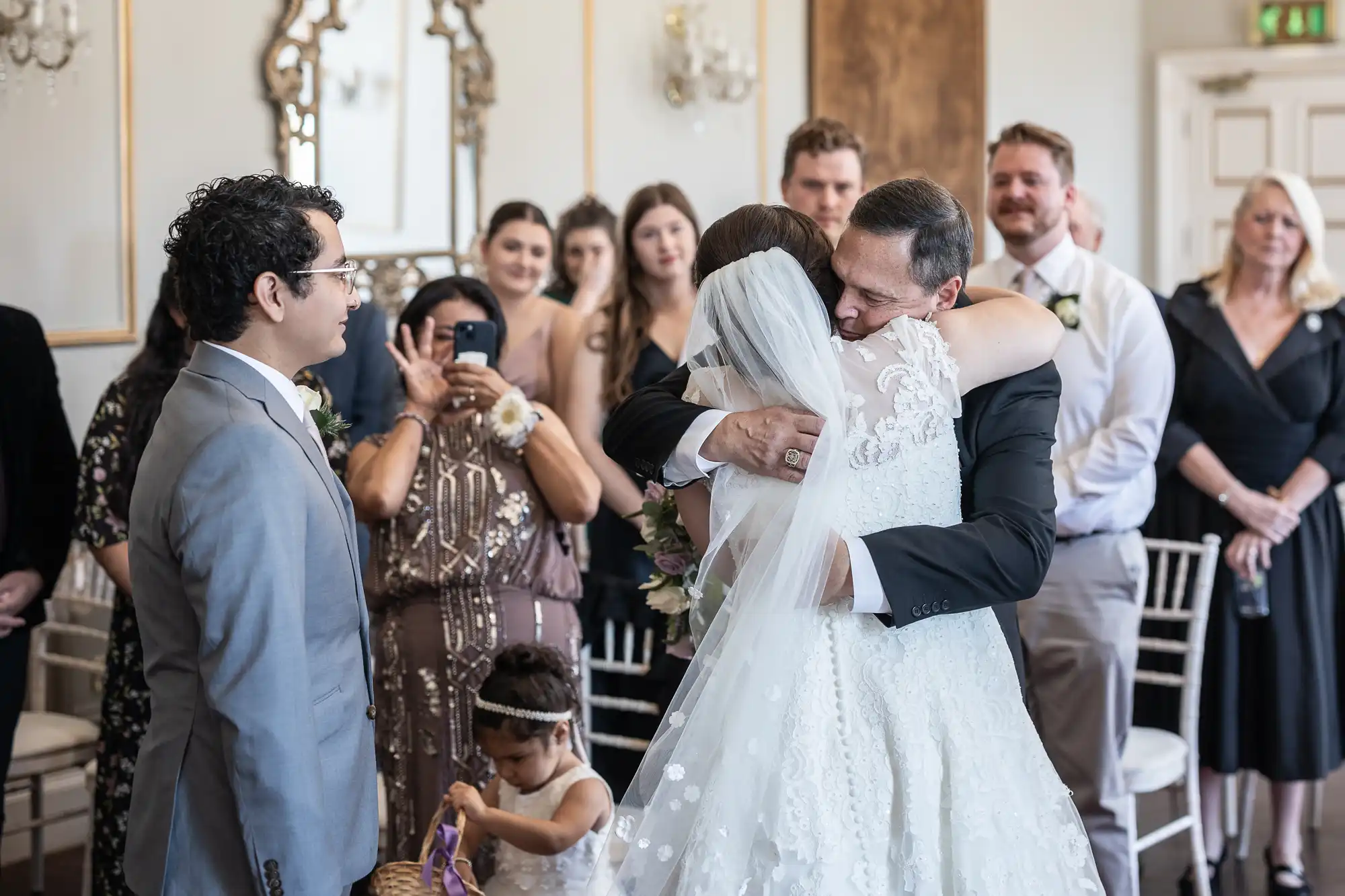 A bride in a white wedding dress hugs a man in a suit, while guests in formal attire look on and a young flower girl stands nearby holding a basket.