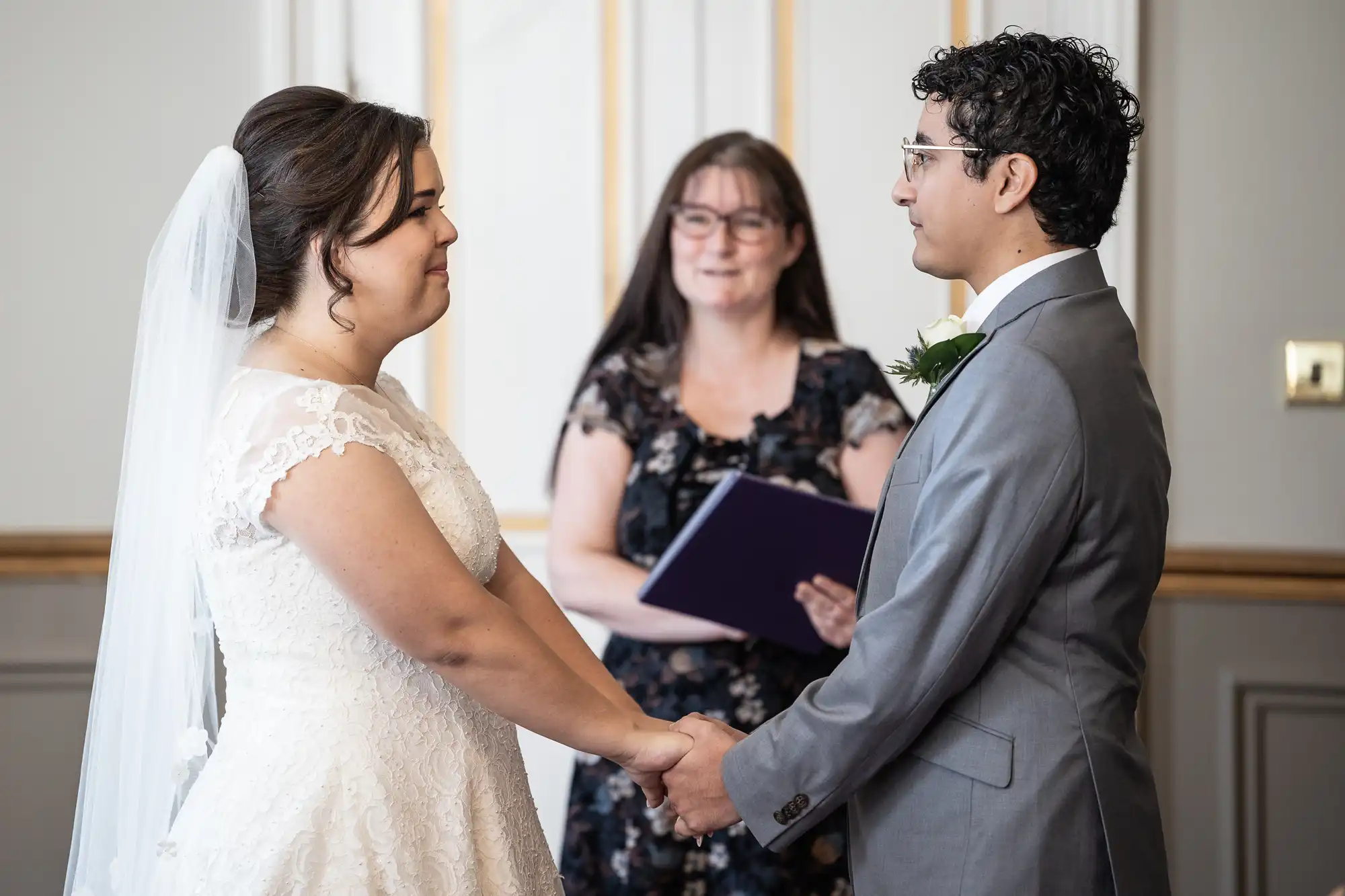 A bride and groom hold hands and face each other during a wedding ceremony while an officiant stands behind them. The bride wears a white dress and veil; the groom is in a gray suit.