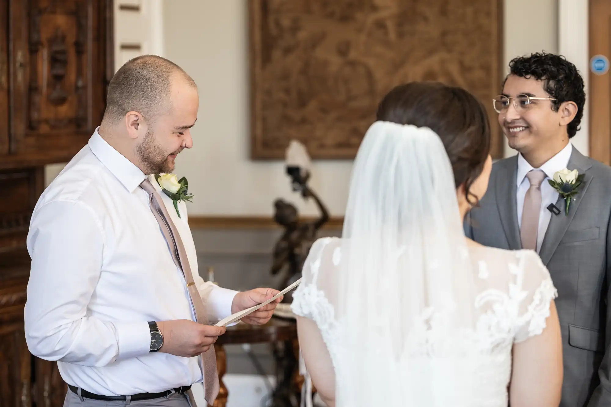 A man in a suit is reading from a paper, while another man in a suit and a woman in a wedding dress stand in front, smiling. They are in an indoor setting with wooden furnishings.