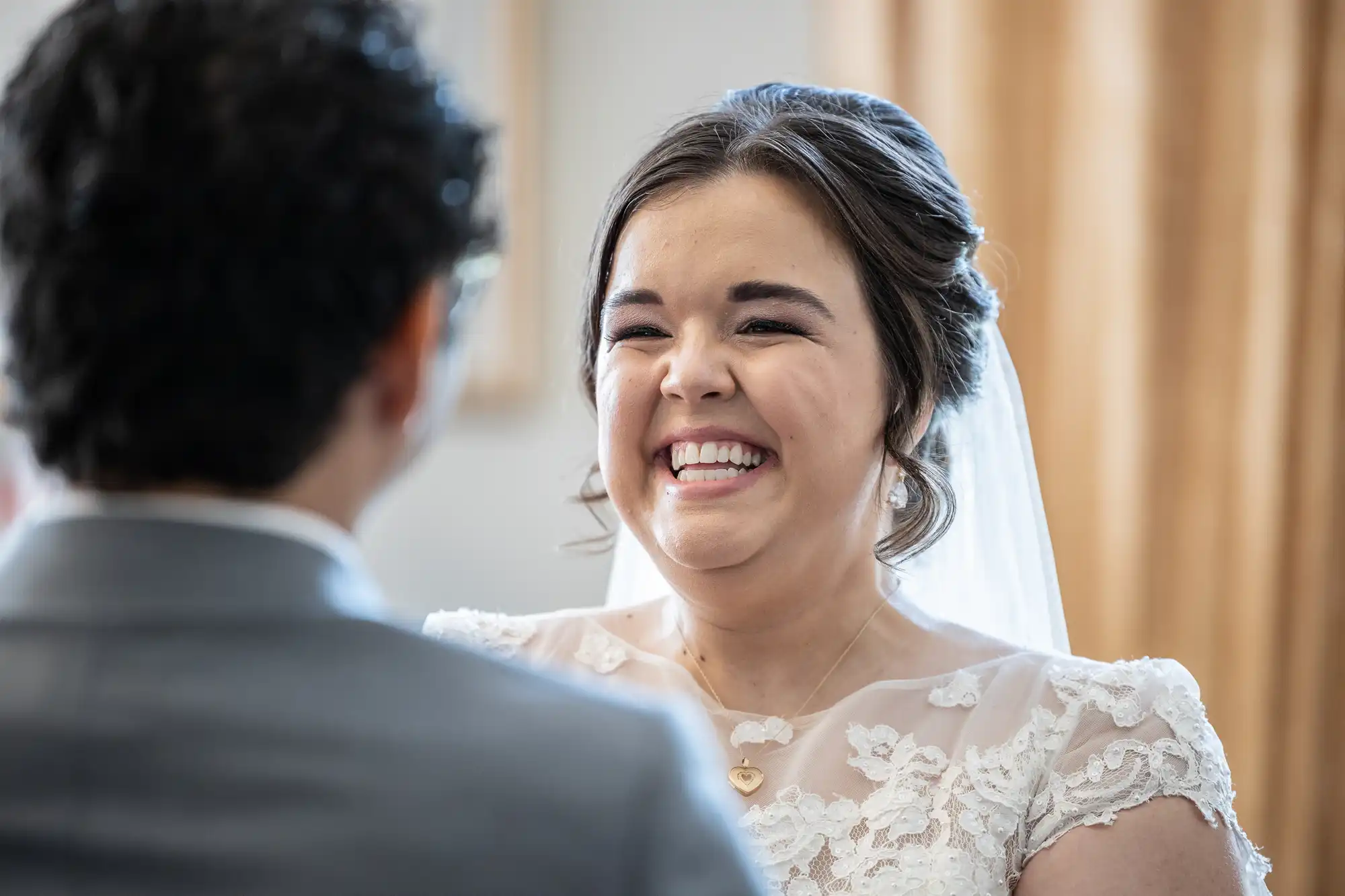 A bride wearing a lace dress and veil smiles widely at a person in a gray suit during a wedding ceremony.