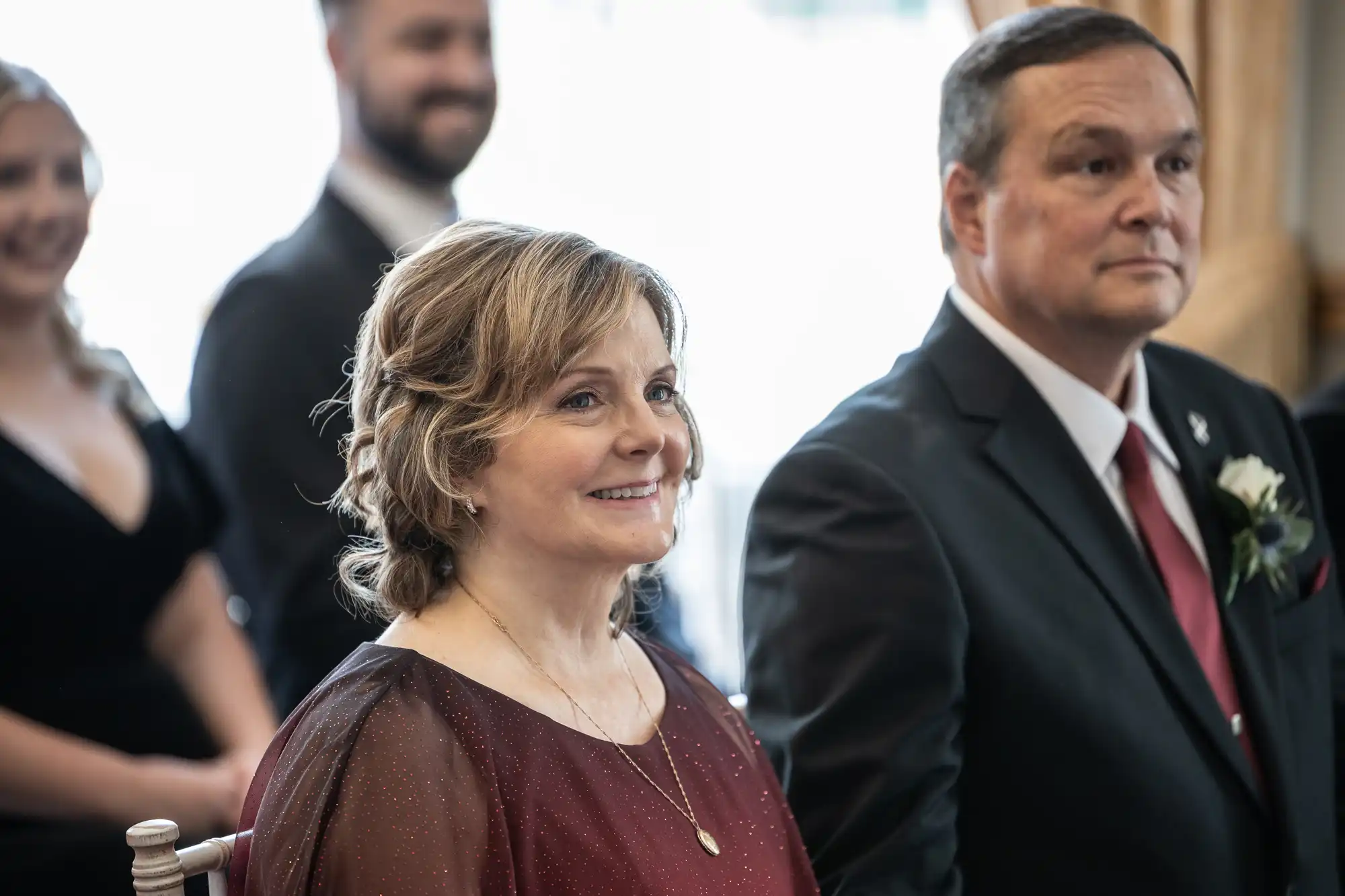 A woman in a maroon dress and a man in a dark suit sit indoors, with people in the background.