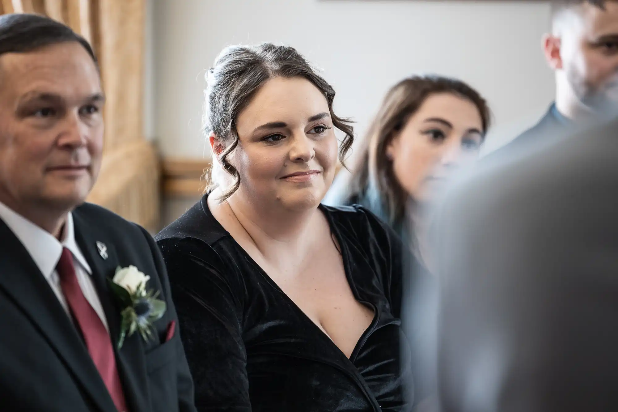 A group of people attend a formal event. The focus is on a woman with brown hair wearing a black dress, sitting among others, listening attentively.