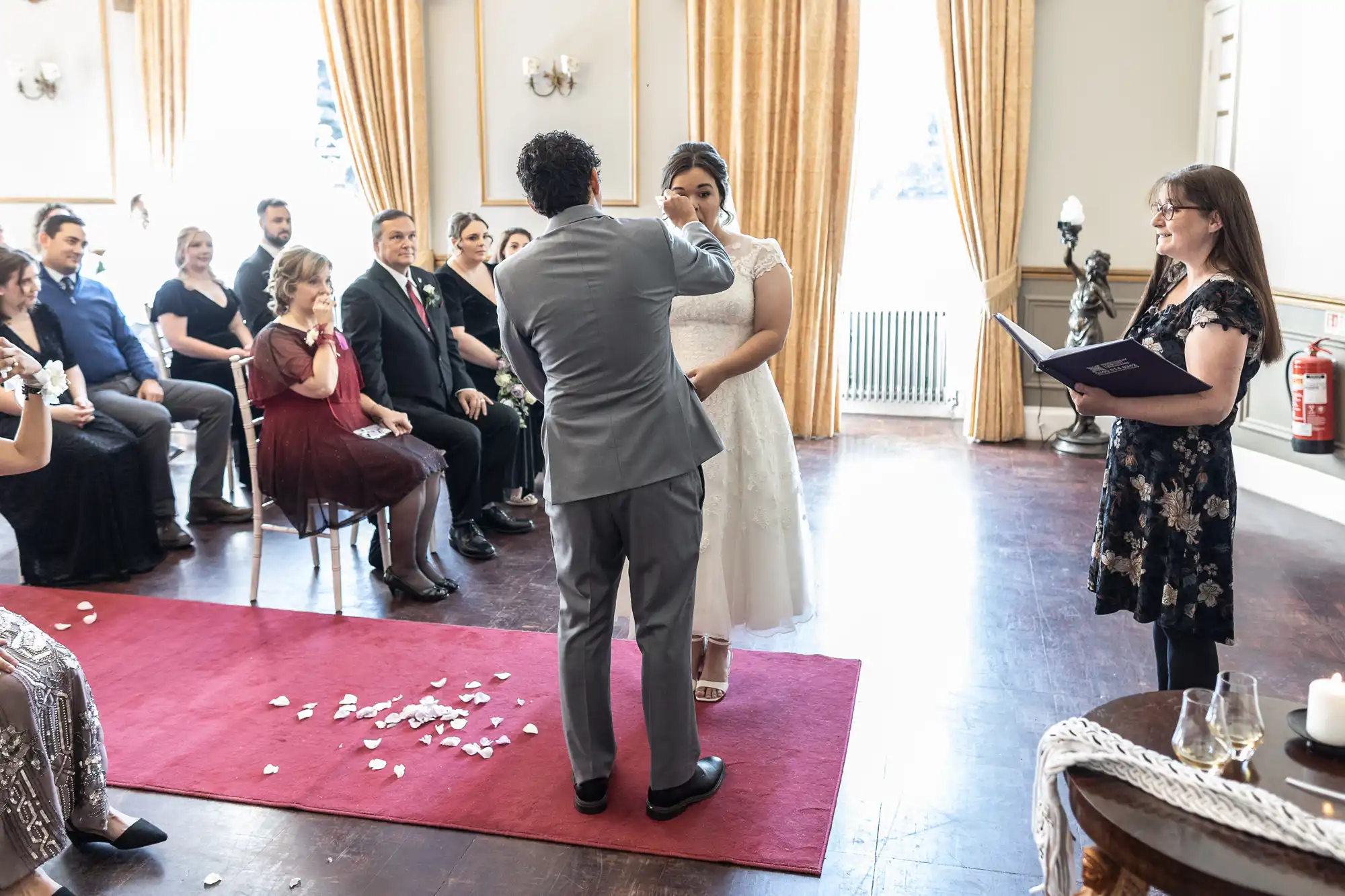 A bride and groom stand in front of guests during a wedding ceremony. The groom is placing a ring on the bride's hand. An officiant holding a book stands nearby. Rose petals are scattered on the floor.