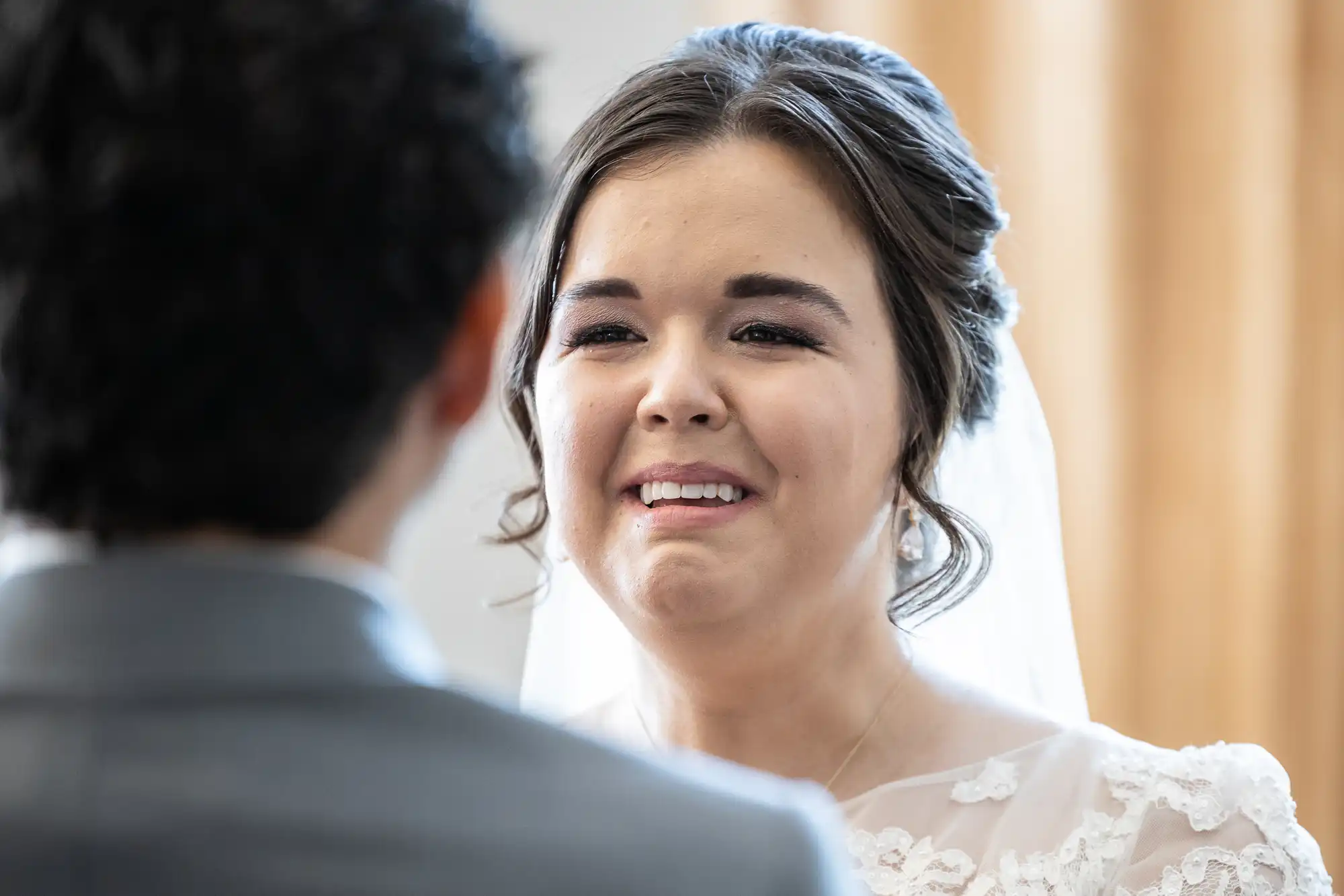 A bride in a white dress and veil smiles at a groom in a gray suit during a wedding ceremony.