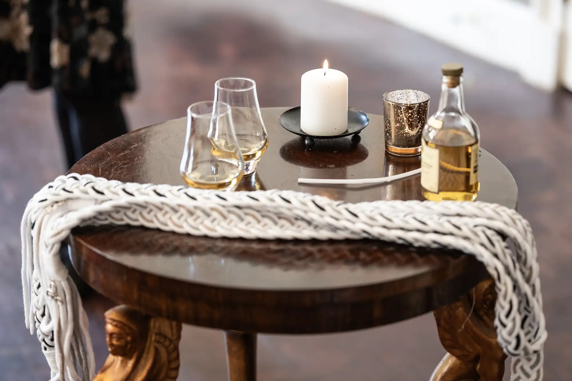 A round wooden table with two glasses of whiskey, a bottle of whiskey, a lit candle, a decorative cup, and a knitted scarf draped across it.