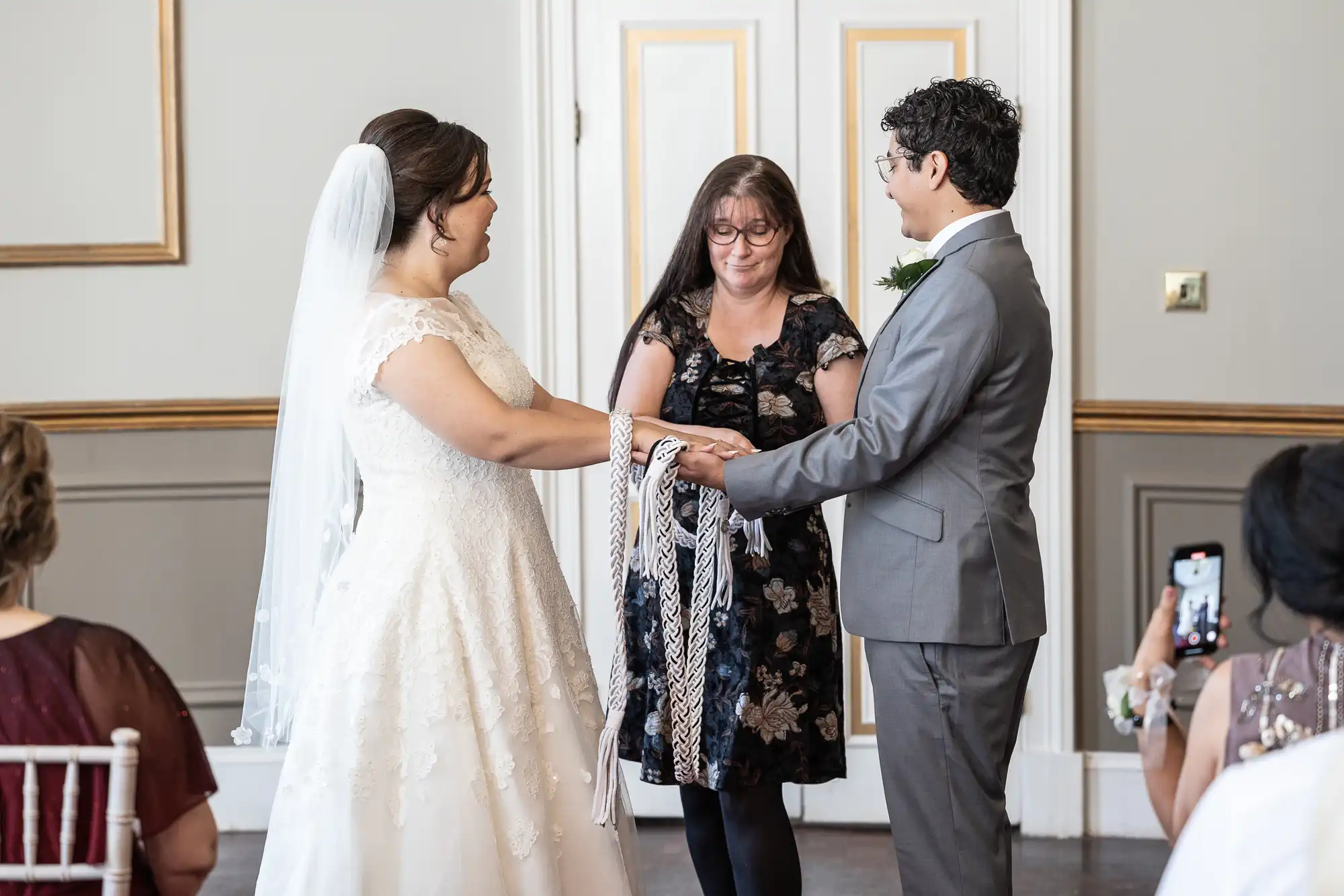 A bride and groom stand facing each other, holding hands joined by a cloth, with an officiant in between them. Guests are seated nearby, one capturing the moment on their phone.