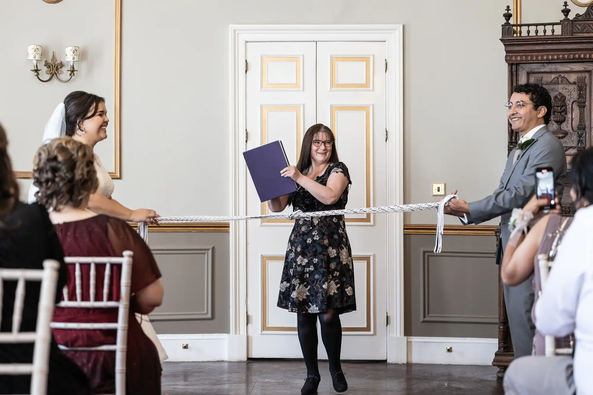 A woman holds a book while a couple, in formal attire, holds a ribbon at a ceremony. Attendees are seated and observing the event.