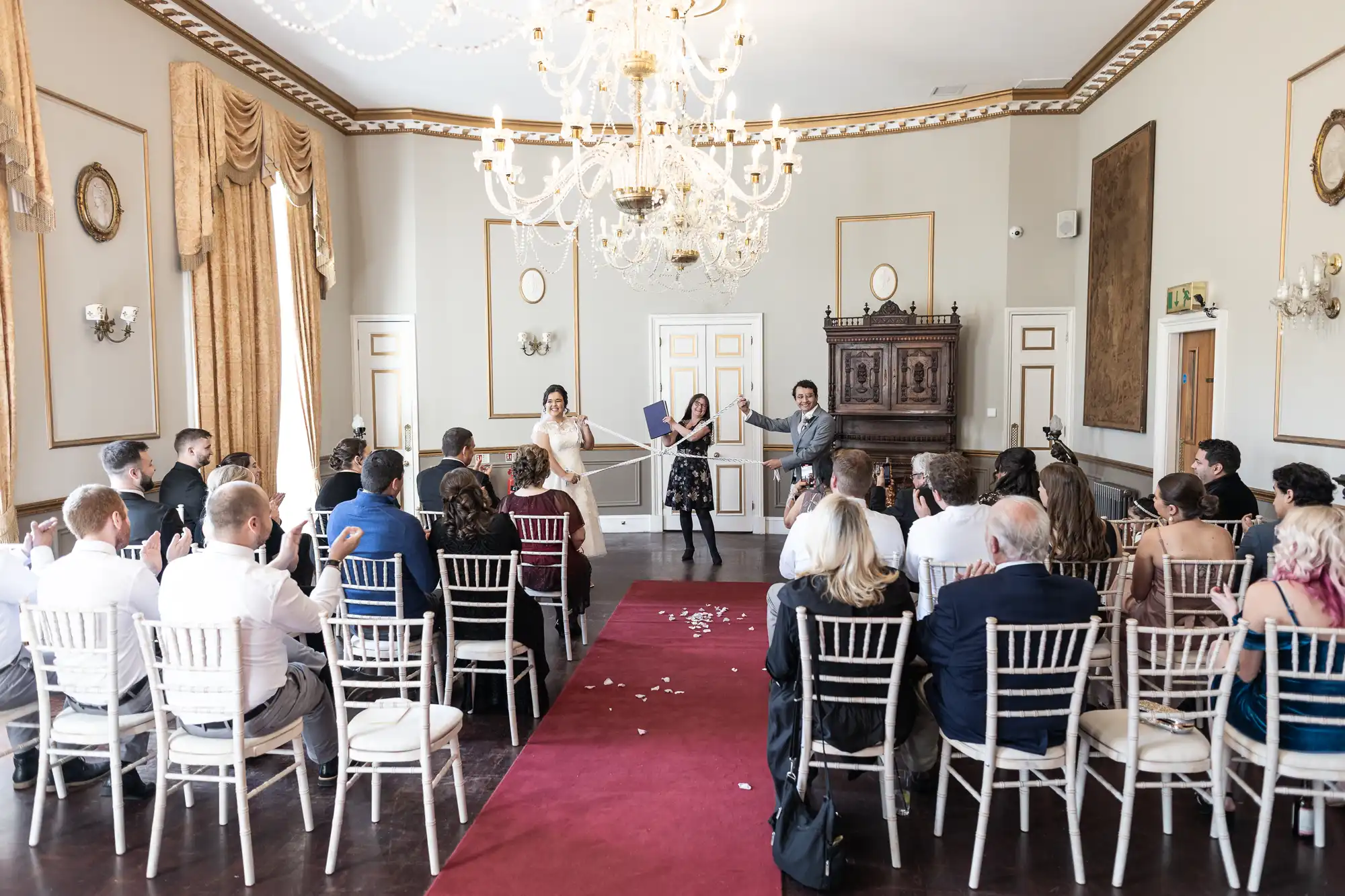 A group of people is seated in a room with elegant decor, attending a ceremony. A red carpet leads to a couple standing at the front, holding a certificate, with a few scattered petals on the floor.