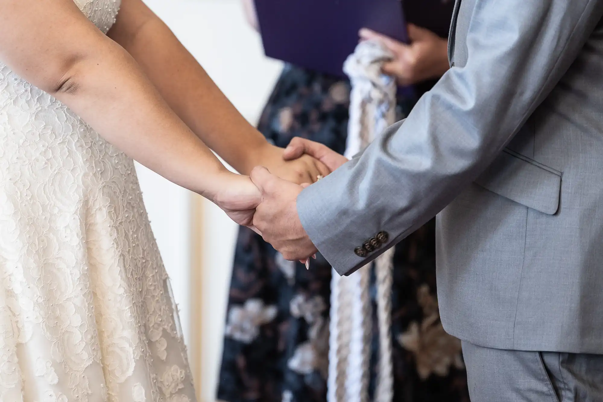 A couple holds hands during a wedding ceremony, with the bride in a lace dress and the groom in a gray suit. A person in the background holds a purple book and a white rope.