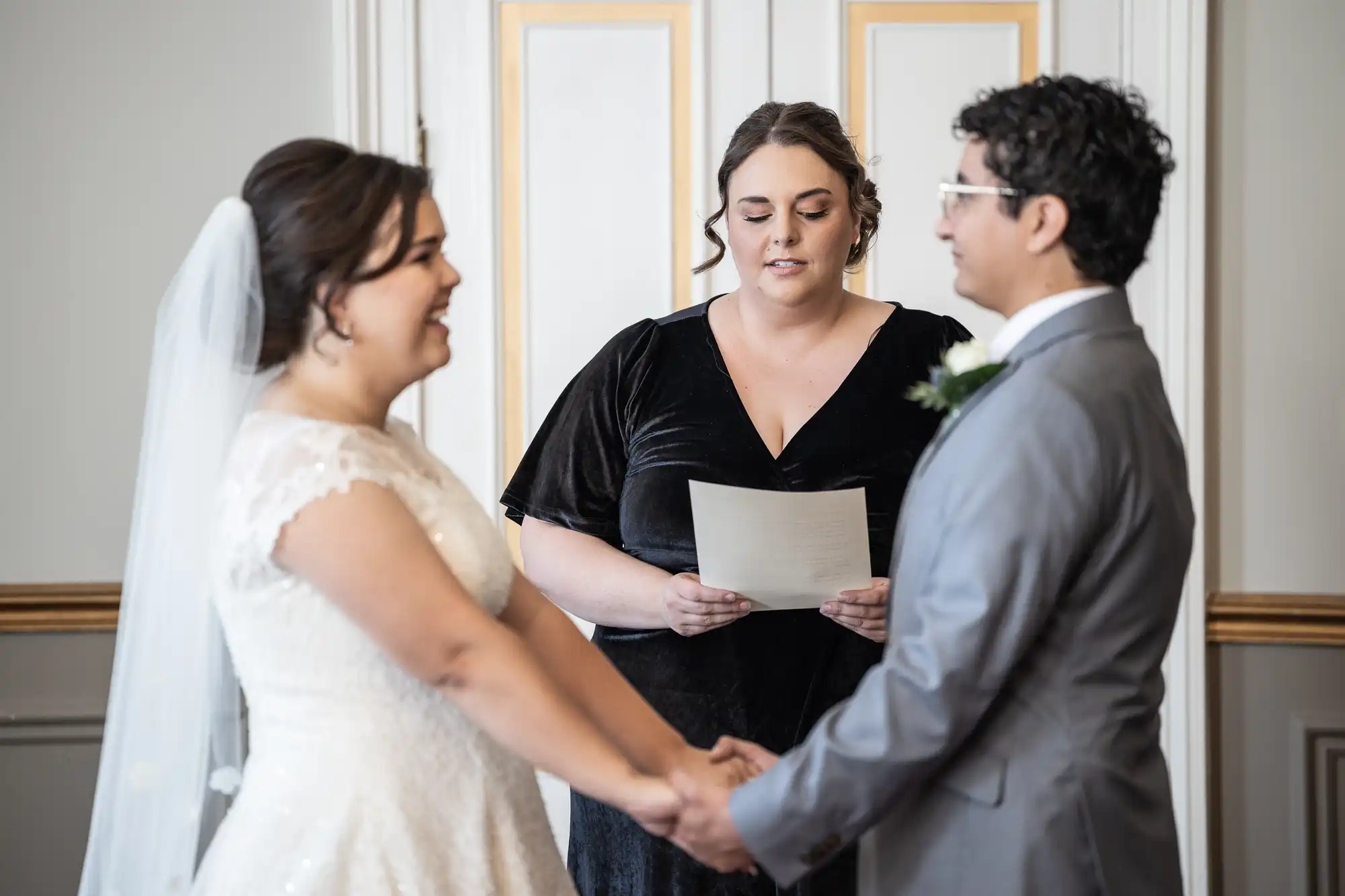 A couple holds hands and smiles at each other during their wedding ceremony, with an officiant reading from a paper in the background.