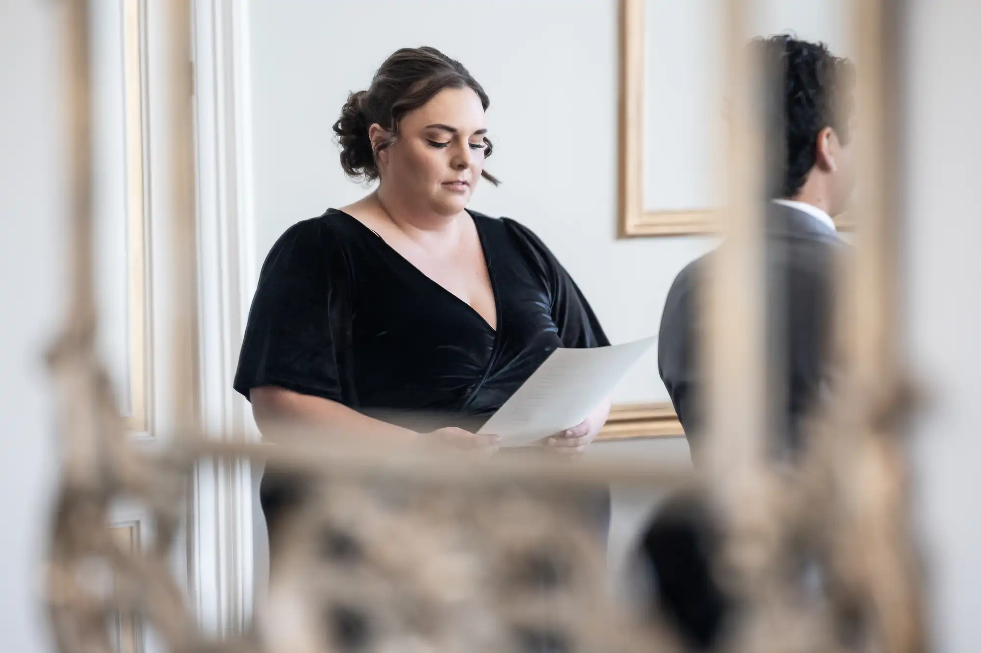 A woman in a black dress is reading from a piece of paper. A man stands beside her, facing away. The background features light-colored walls with decorative frames.