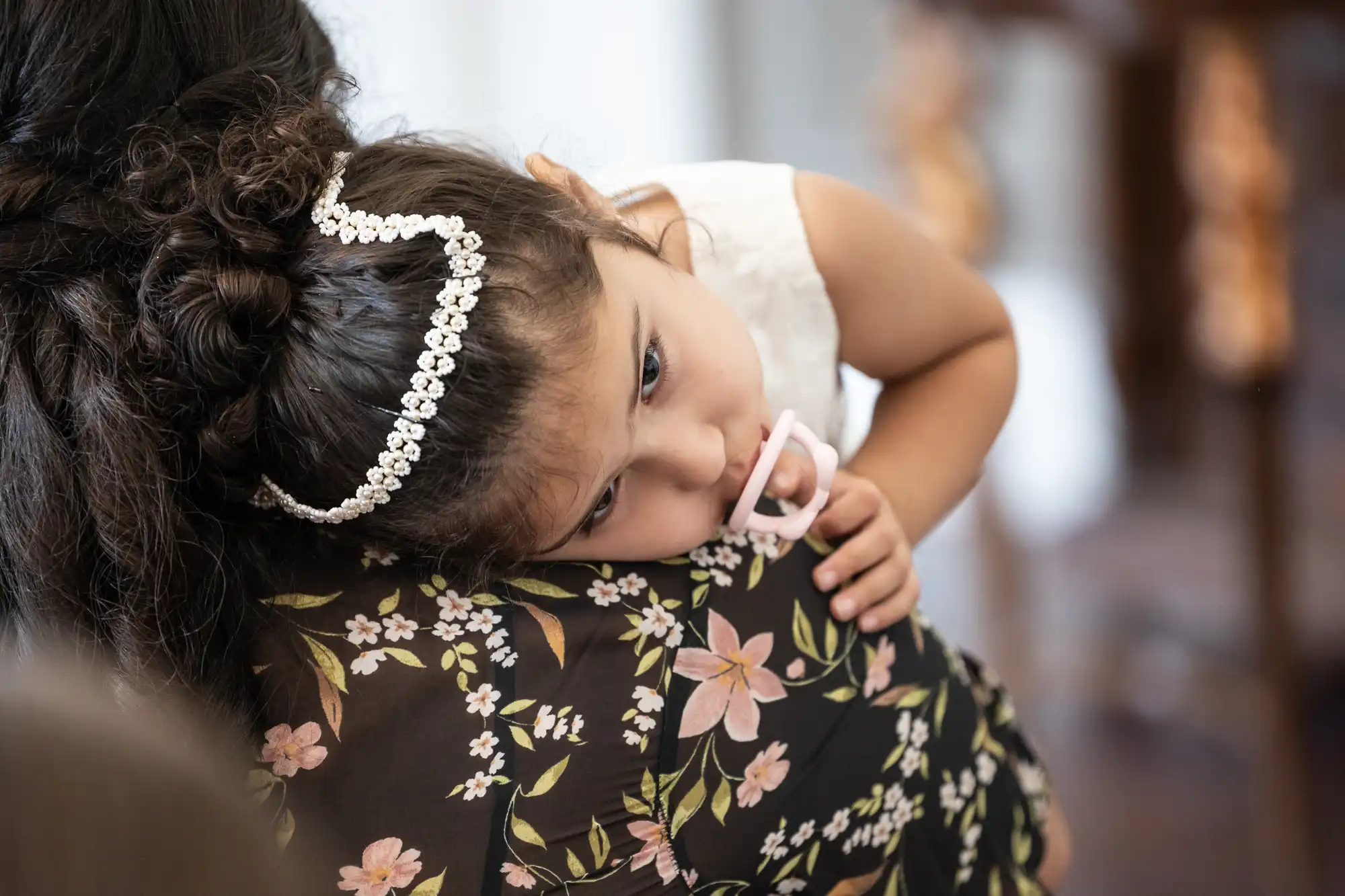 A young child wearing a floral headband rests her head on an adult's shoulder. The adult is wearing a black floral top.