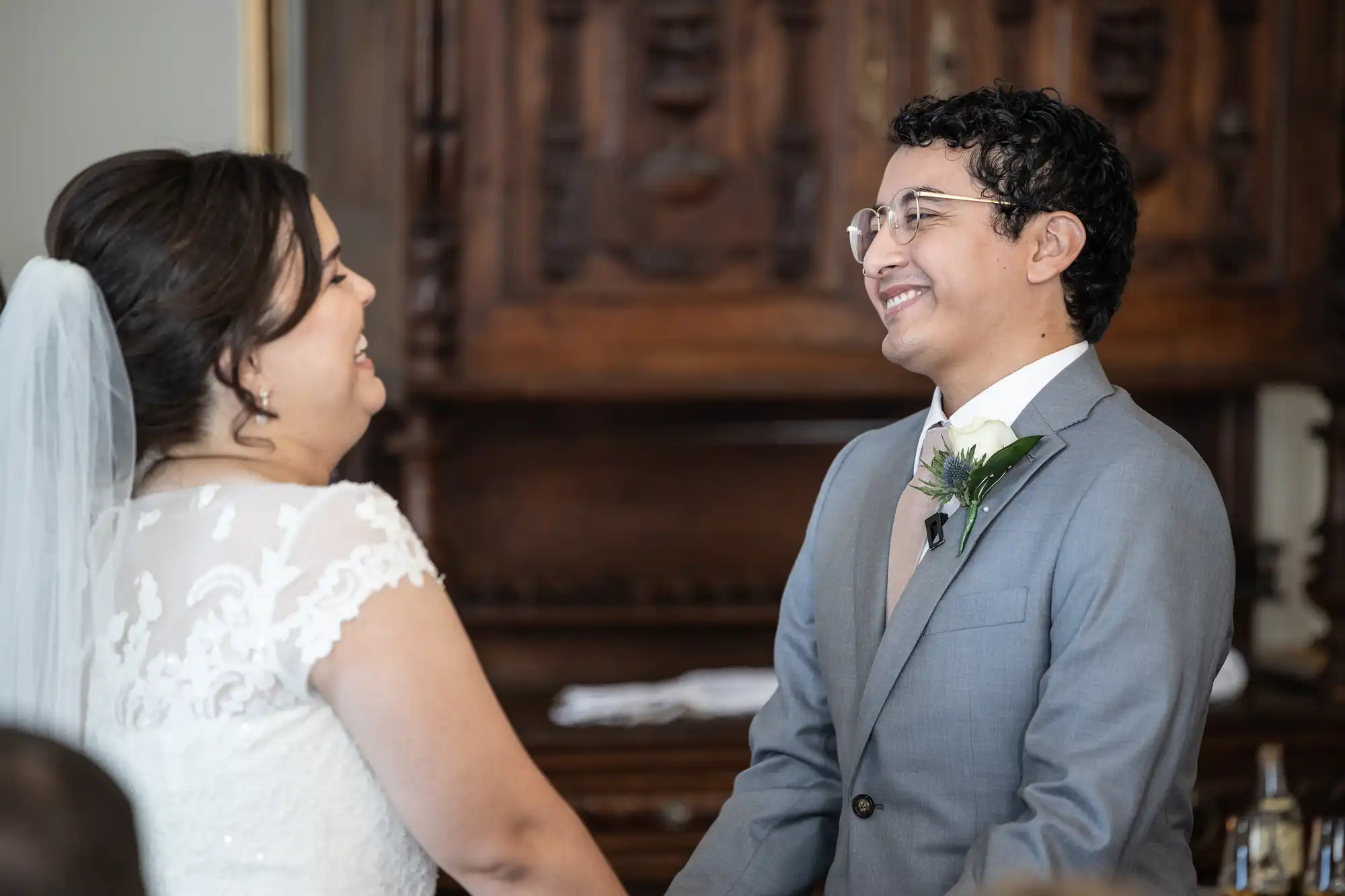 A bride and groom smile at each other during their wedding ceremony, with the bride wearing a white dress and veil and the groom in a gray suit with a boutonniere.