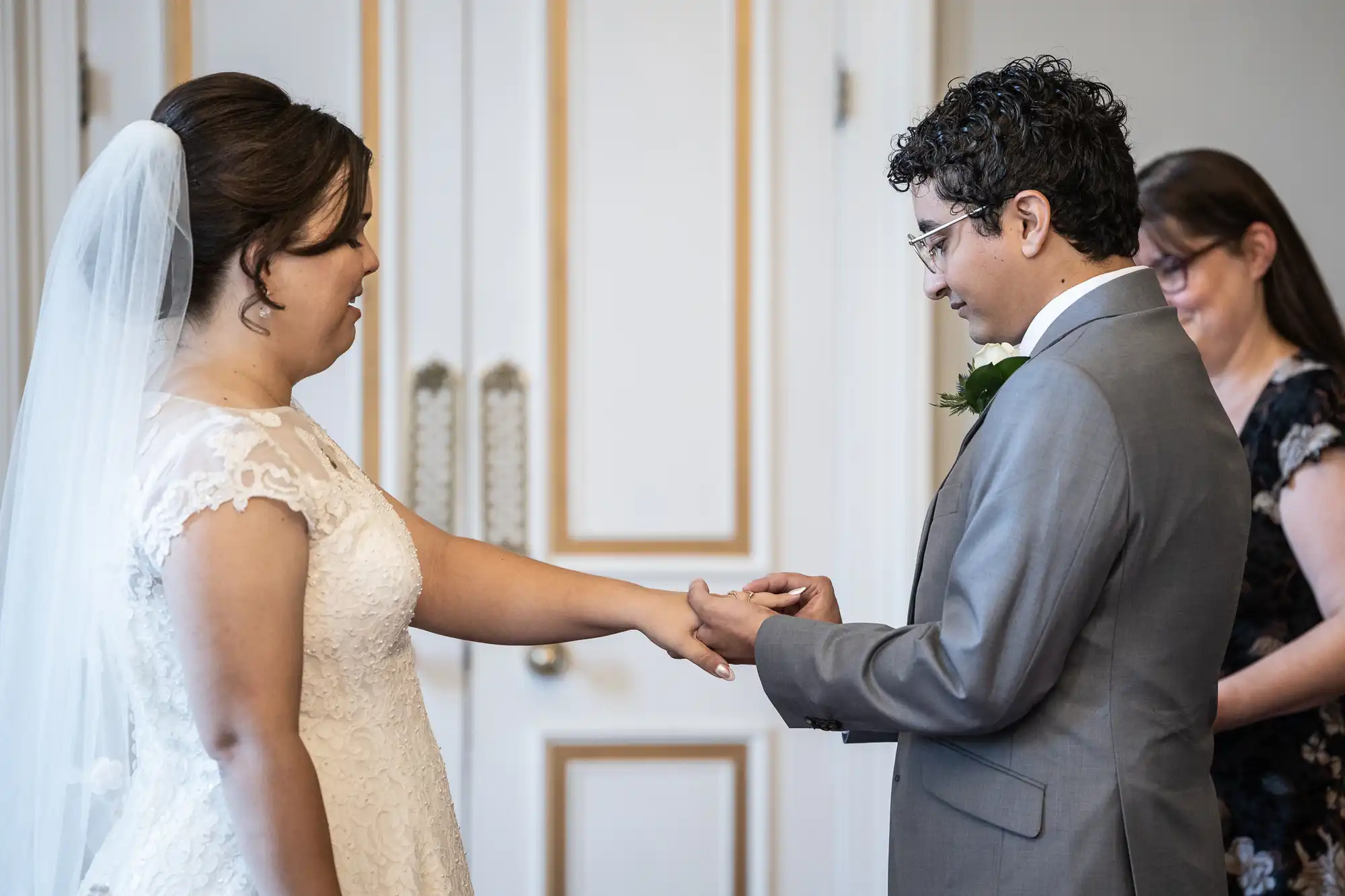 Two people exchange rings during a wedding ceremony, with one wearing a white dress and veil, and the other in a grey suit. A person in the background observes the moment.