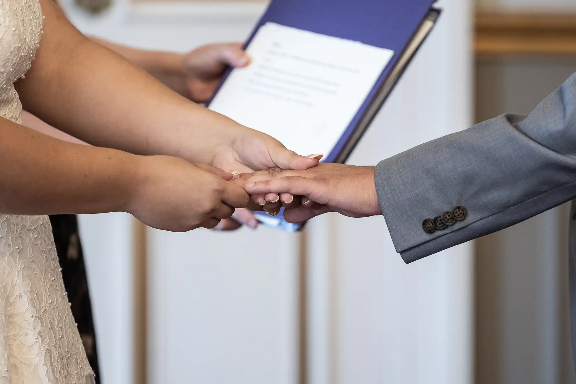 Two people holding hands during a formal event, with one person in a white dress and the other in a light gray suit. A third person is holding a blue folder in the background.