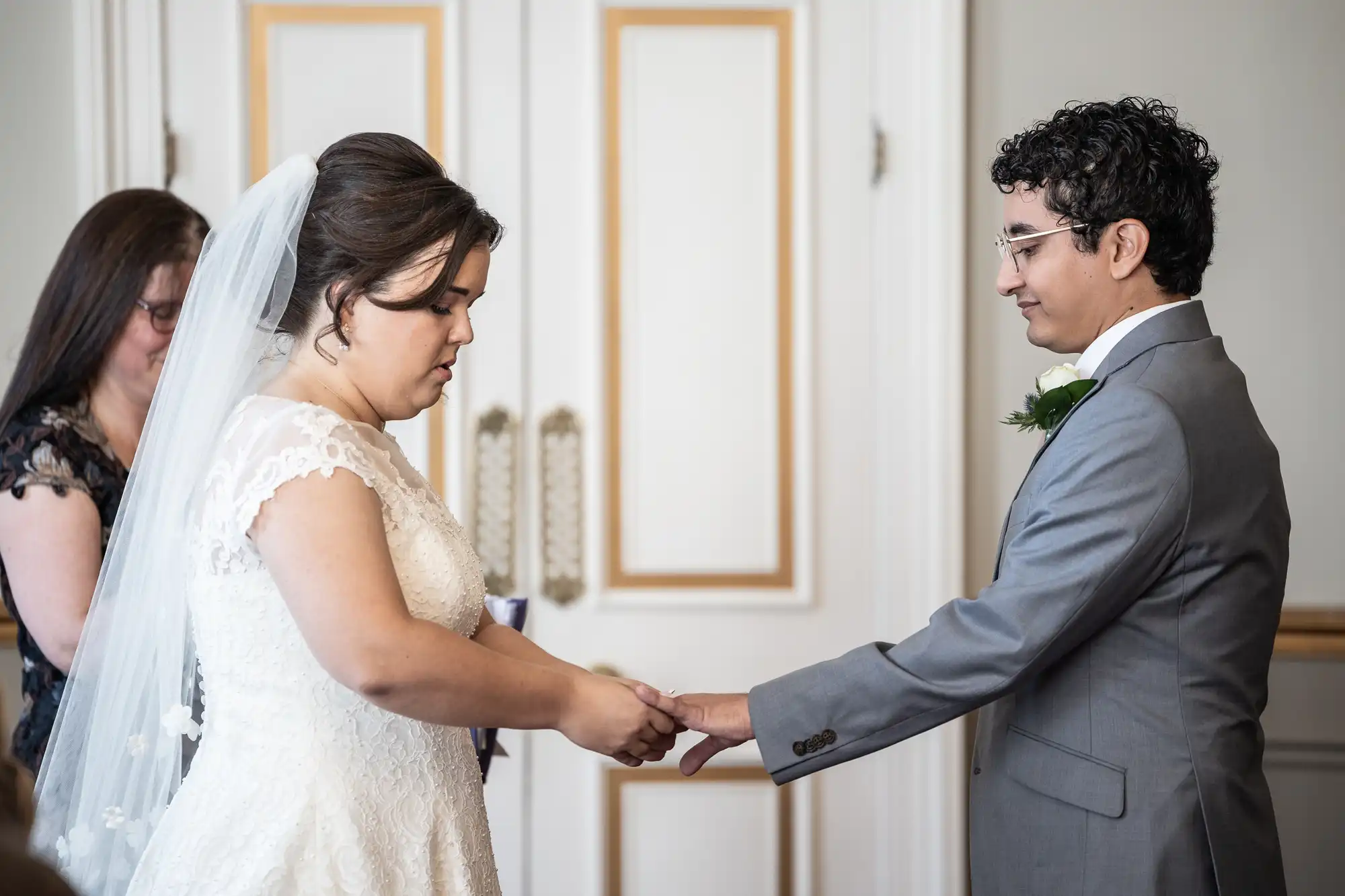 A bride and groom exchange rings during a wedding ceremony. The bride is in a white dress and veil, while the groom wears a gray suit. A woman stands in the background.