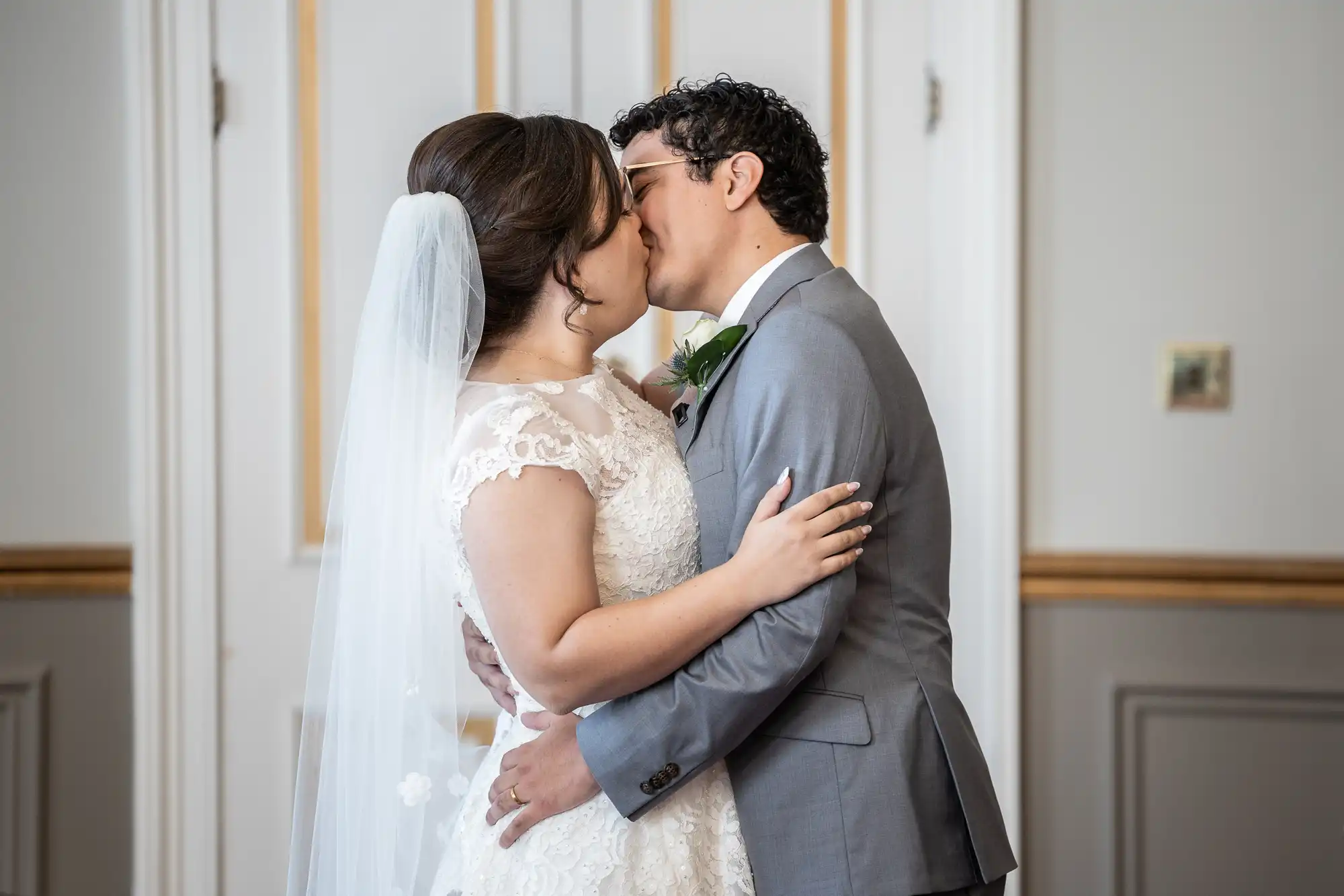 A bride and groom share a kiss during their wedding ceremony in a room with white and gold doors.