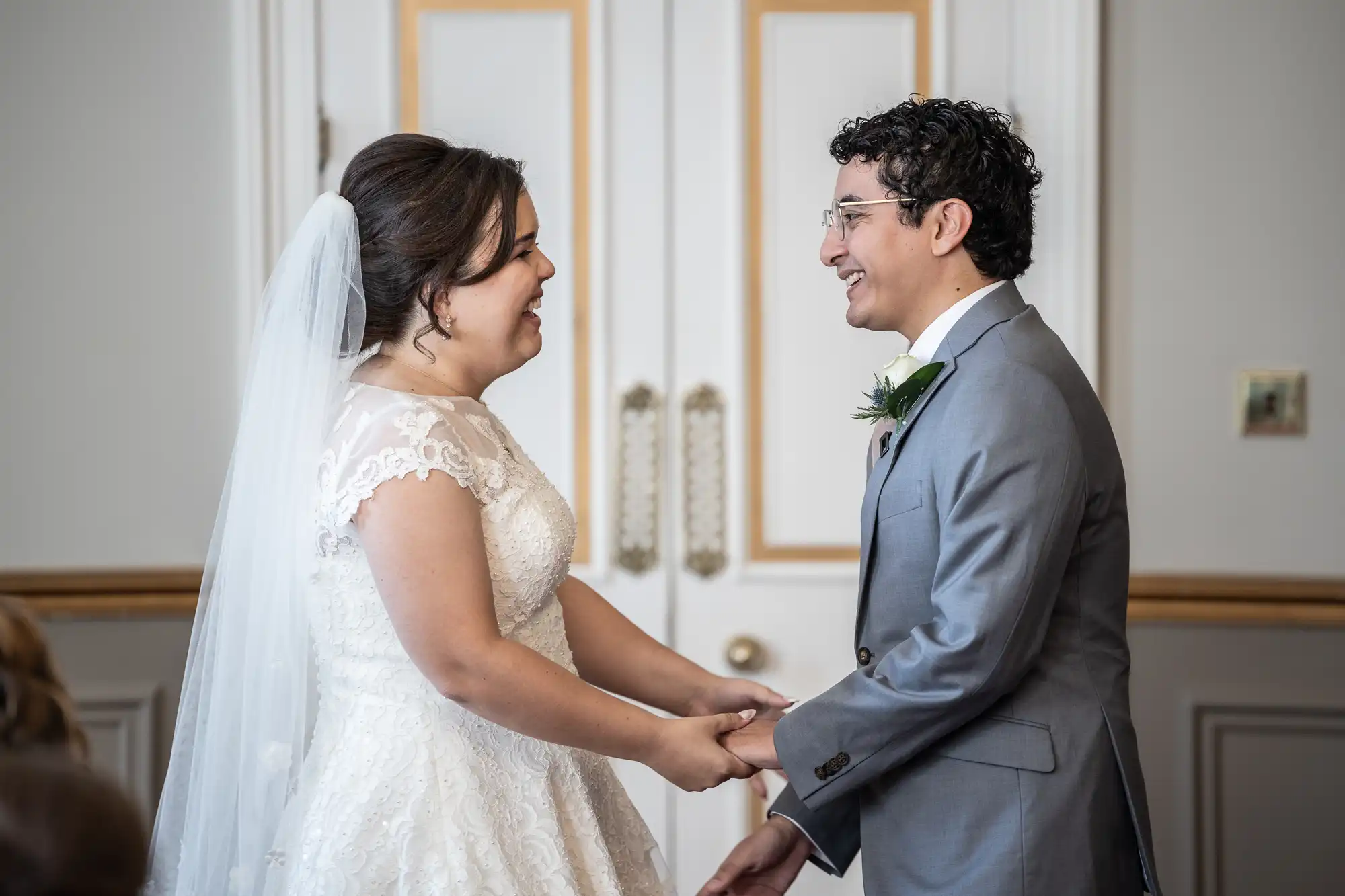 A bride and groom hold hands and smile at each other during their wedding ceremony in front of double doors with gold accents.