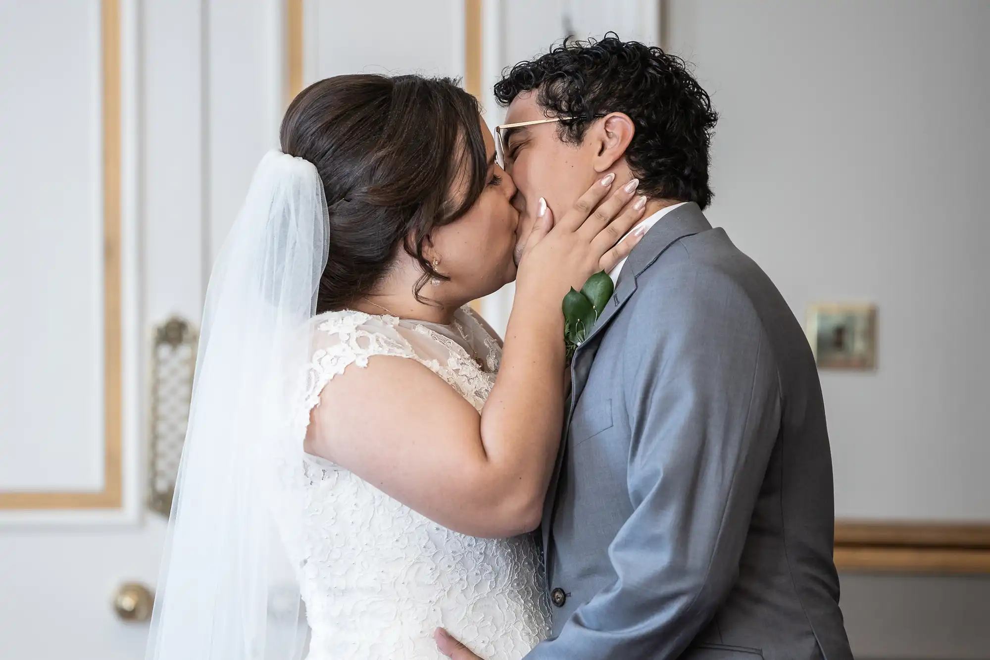 A bride in a white lace dress and veil kisses a groom in a gray suit with glasses.