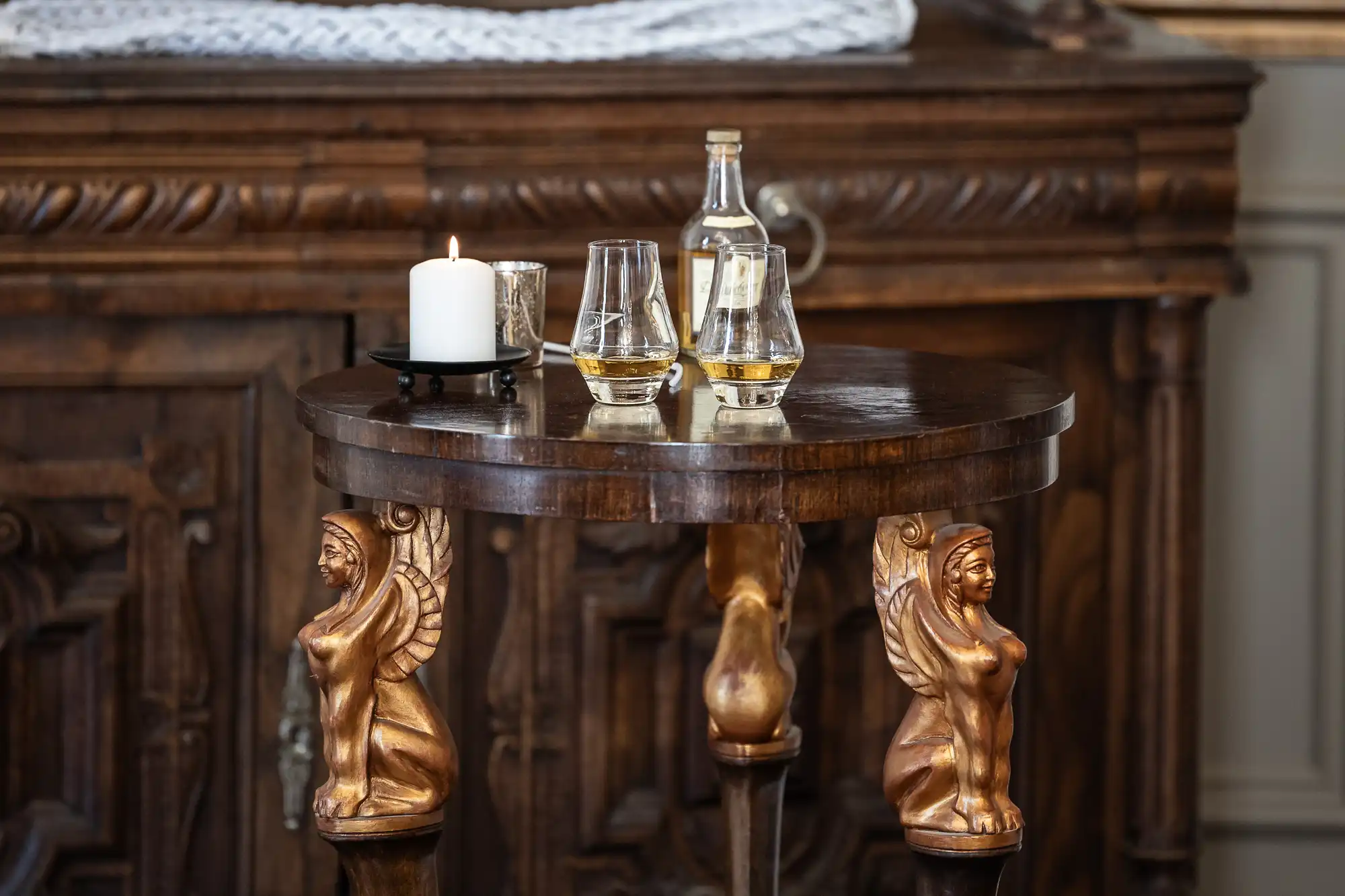 A round wooden table with ornate carvings of mythical figures holds a lit candle, a small glass, and two glass decanters partially filled with amber-colored liquid.