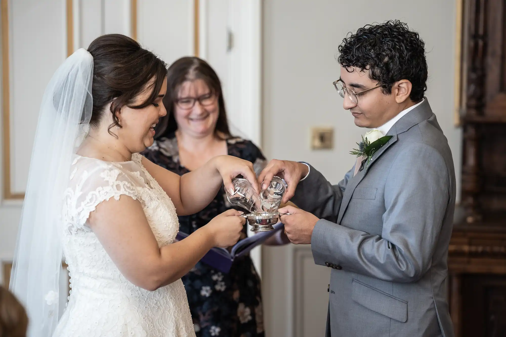 A bride and groom in wedding attire pour sand into a jar during a unity sand ceremony, while a woman stands behind them, smiling.