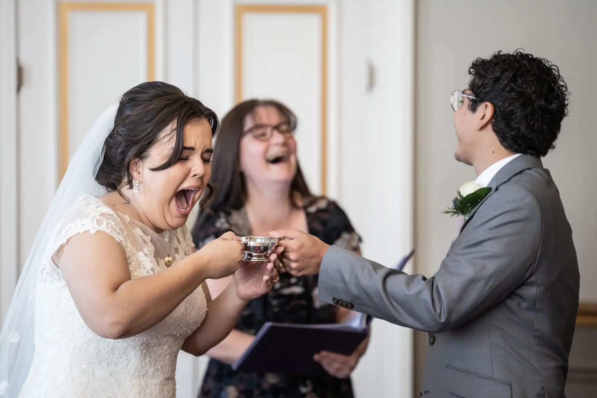 A bride laughs heartily while holding a teacup, and a groom in a suit offers the cup during a wedding ceremony, with an officiant in the background also laughing.