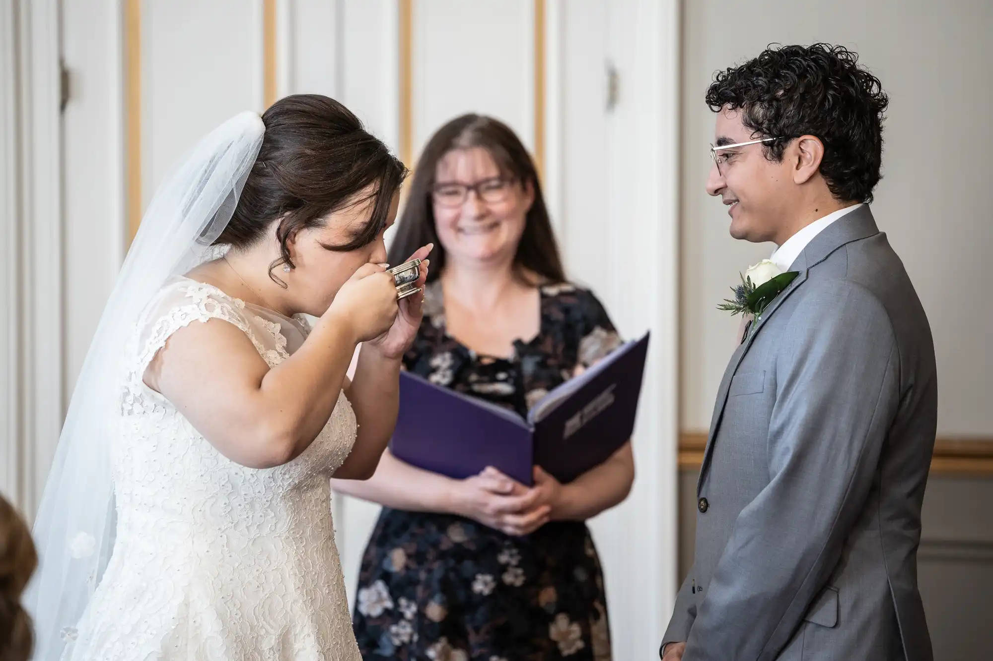 A bride drinks from a small cup while a groom smiles during a wedding ceremony. An officiant holding a folder stands in the background, smiling.