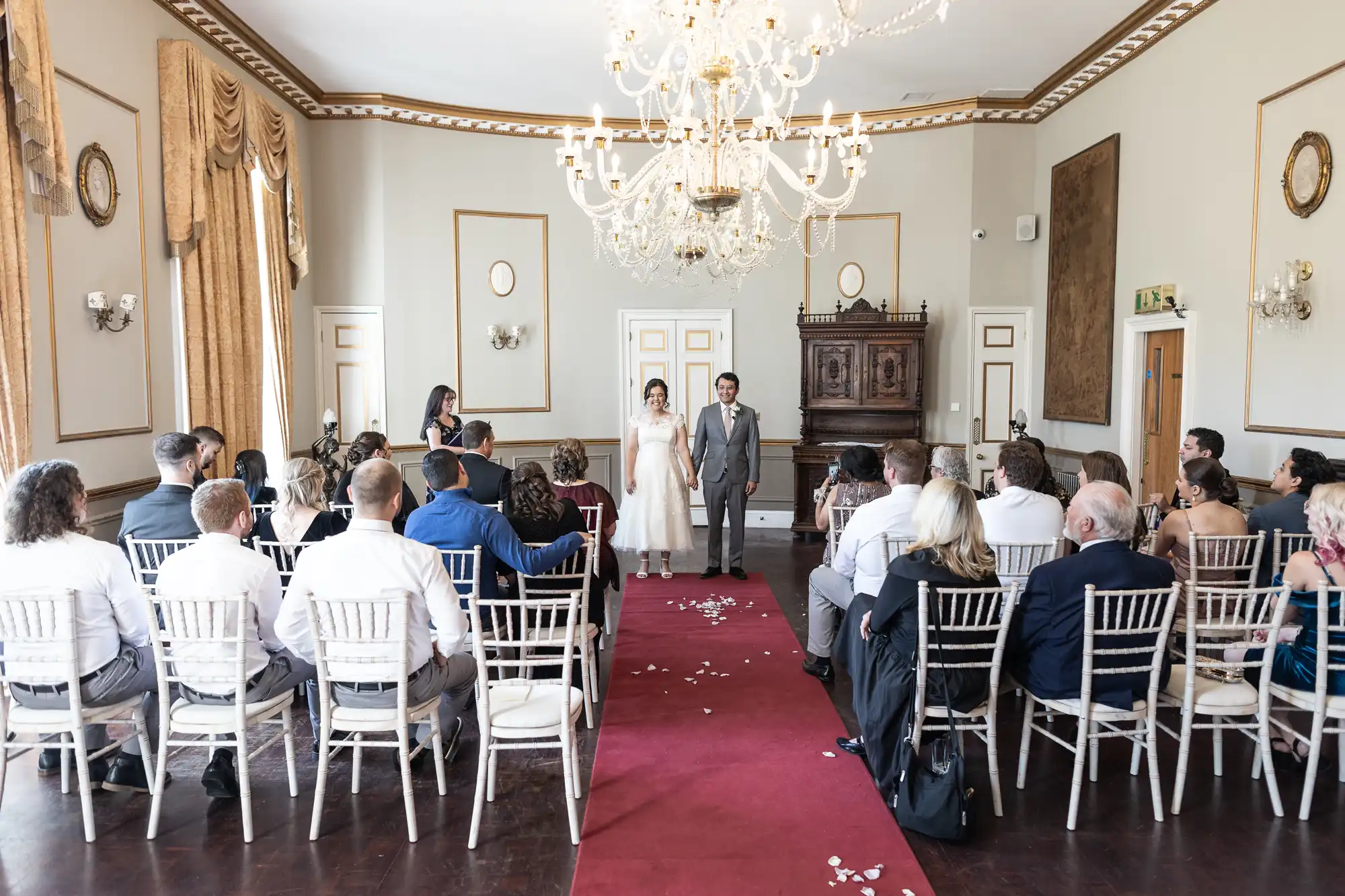 A bride and groom stand at the front of a decorated room, facing seated guests. The room features chandeliers, gold drapes, and an ornate wooden piece at the back.