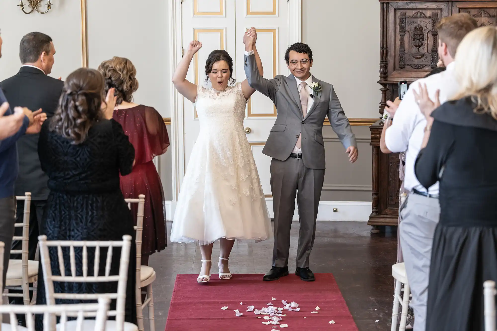 A newlywed couple stands hand in hand, raising their arms in celebration at the end of the aisle, surrounded by clapping guests in a formal room with white chairs and a red carpet.