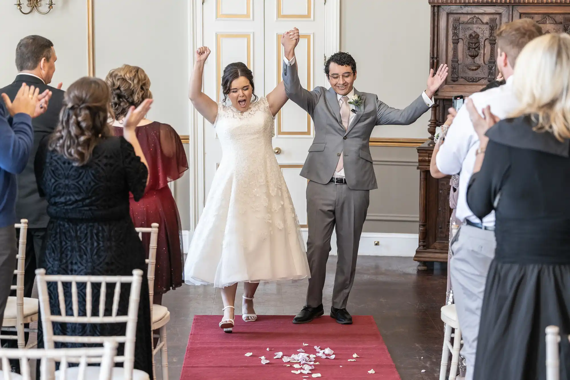 Newlyweds walk down the aisle, holding hands and celebrating as guests applaud. The bride wears a white dress and the groom wears a gray suit.