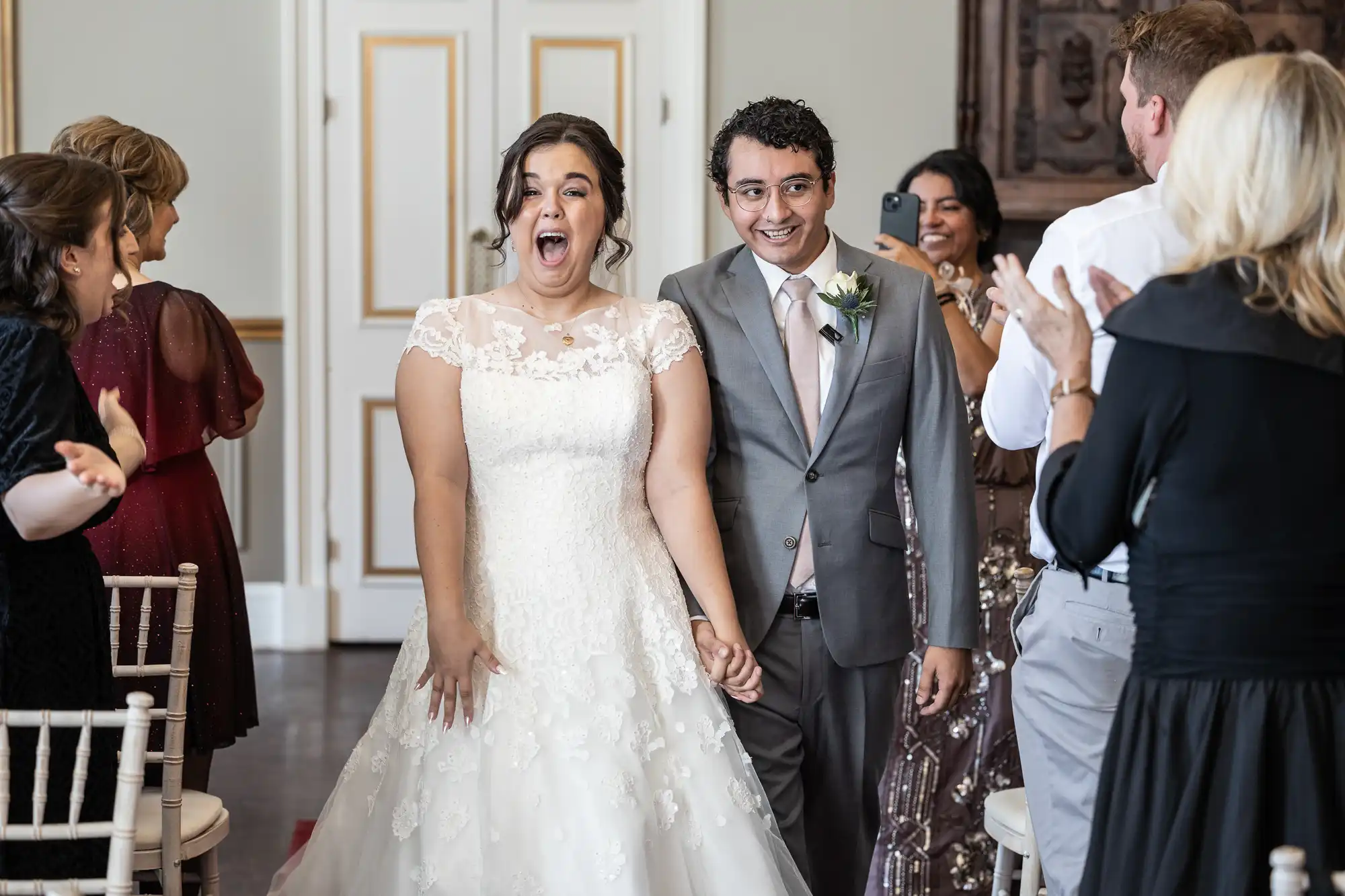 A bride in a white dress and groom in a gray suit, both smiling and holding hands, enter a room while applauding guests look on.