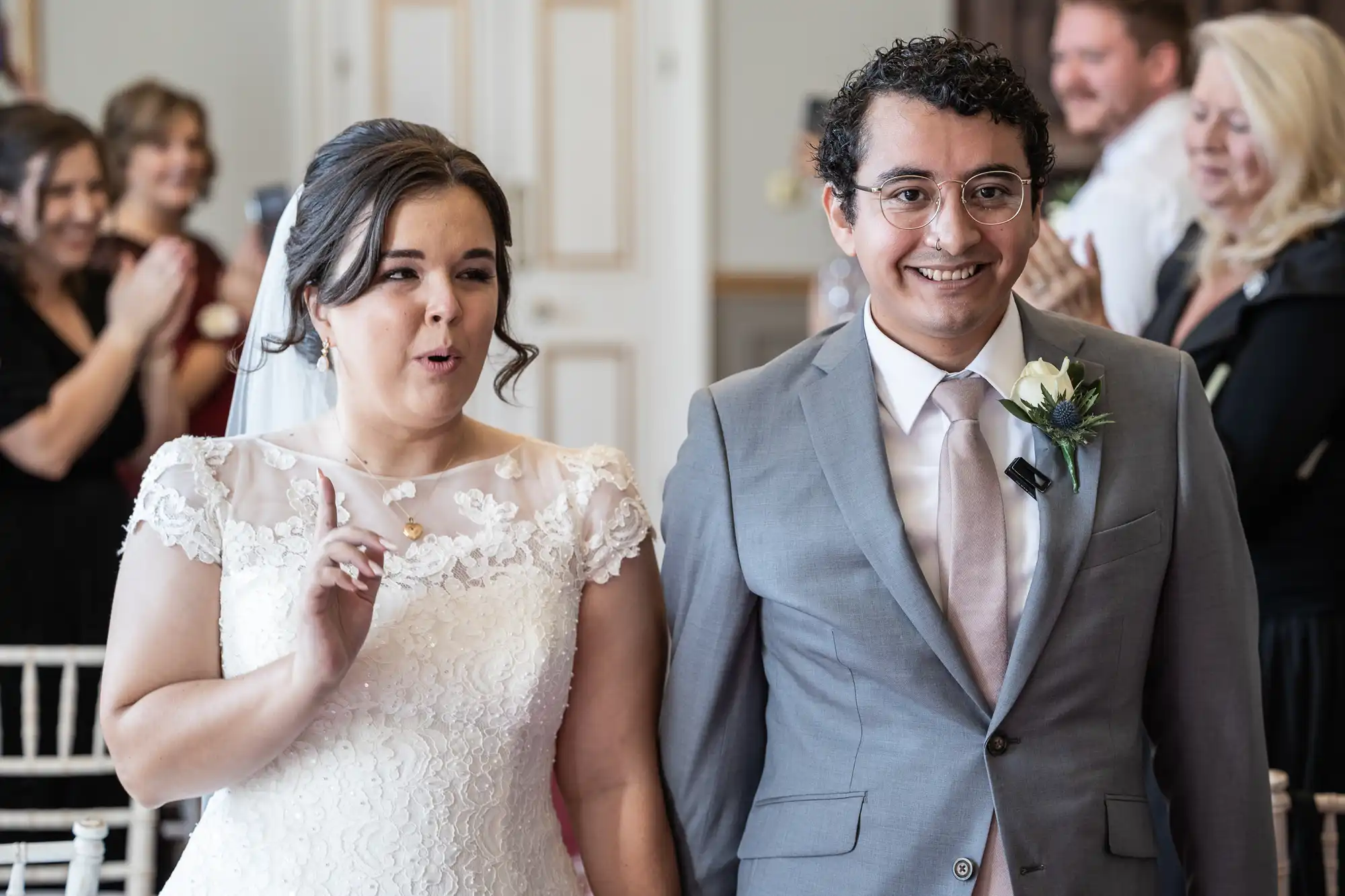 A bride and groom in wedding attire are walking down an aisle, with guests clapping in the background. The bride has her hand raised and is making a gesture, while the groom is smiling.