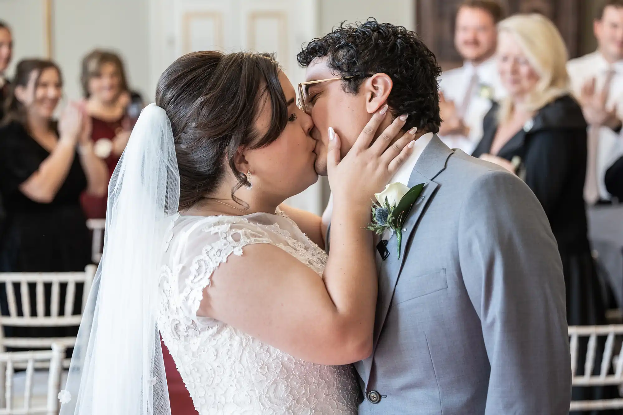 A bride and groom kiss at their wedding ceremony, with guests clapping in the background. The bride is wearing a white dress and veil, and the groom is in a gray suit with a boutonnière.