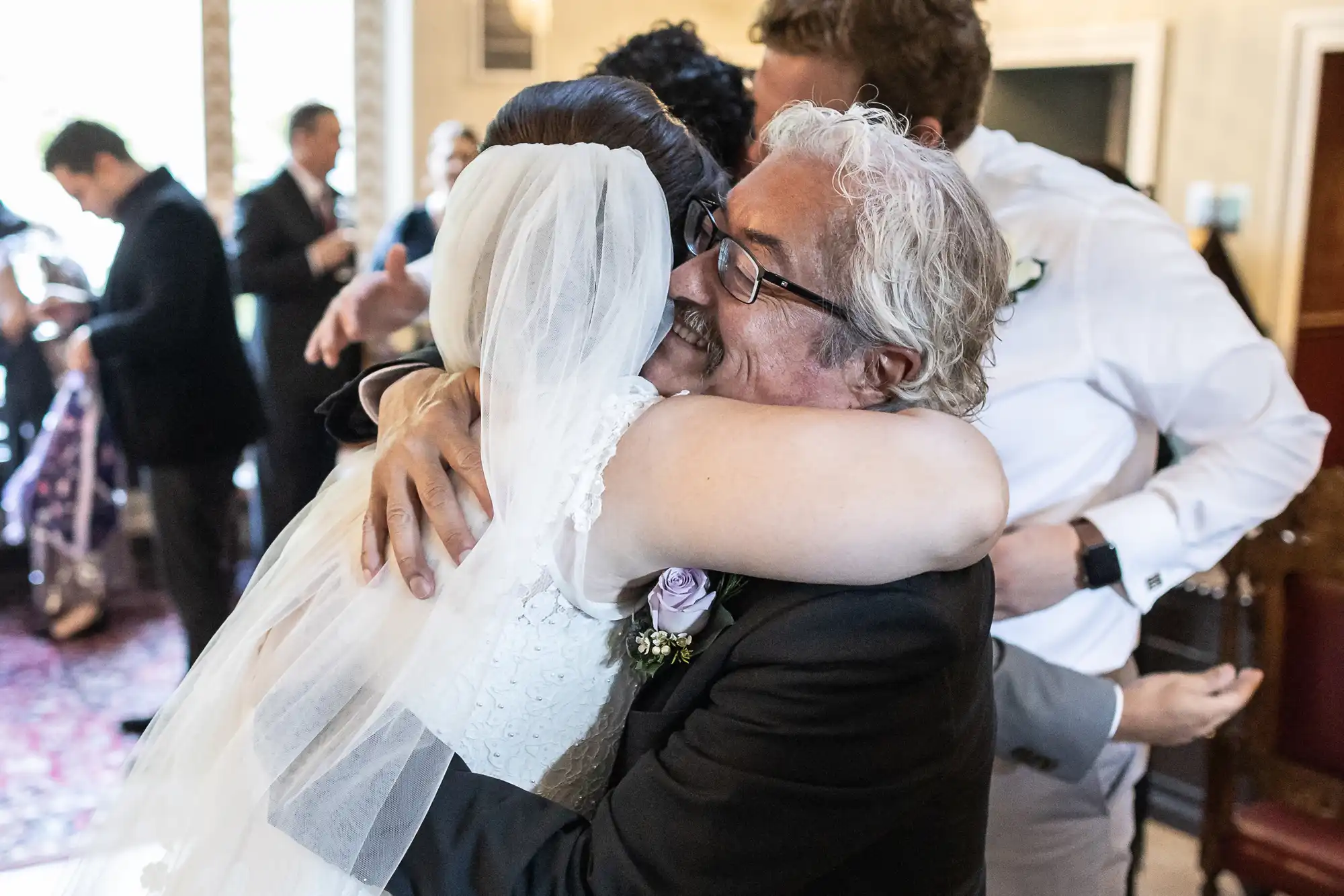 Bride and groom hugging an older man in a room filled with people. The bride is wearing a veil and white dress; the man has white hair and glasses. Other guests are visible in the background.