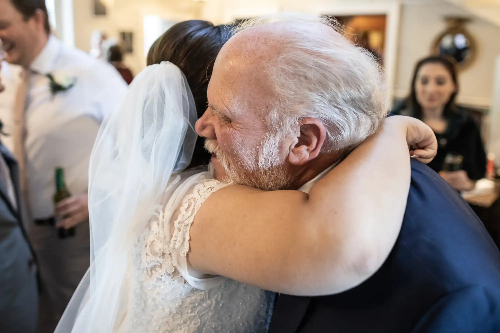 A bride in a wedding dress and veil hugs an older man with white hair and a beard, smiling warmly, in a lively indoor setting.