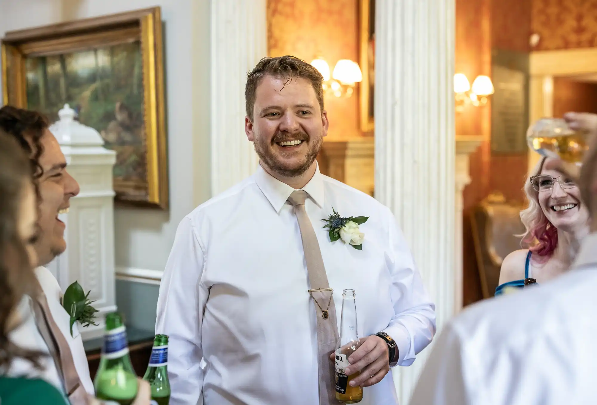 A man in a white shirt and tie holds a drink and smiles while engaging with a group of people. He has a white flower pinned to his shirt. Others in the group are also holding drinks and smiling.