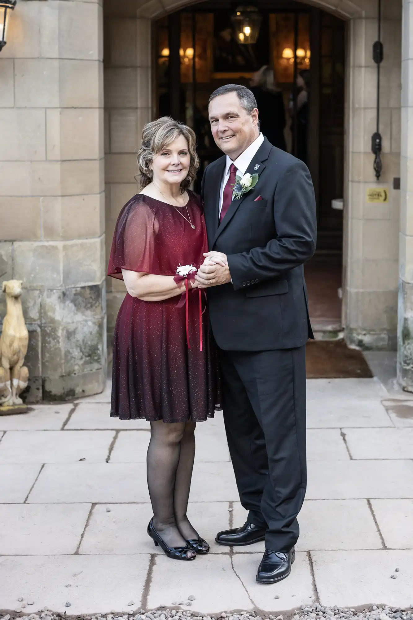 A man in a suit and a woman in a red dress stand smiling, holding hands in front of a stone building entrance.