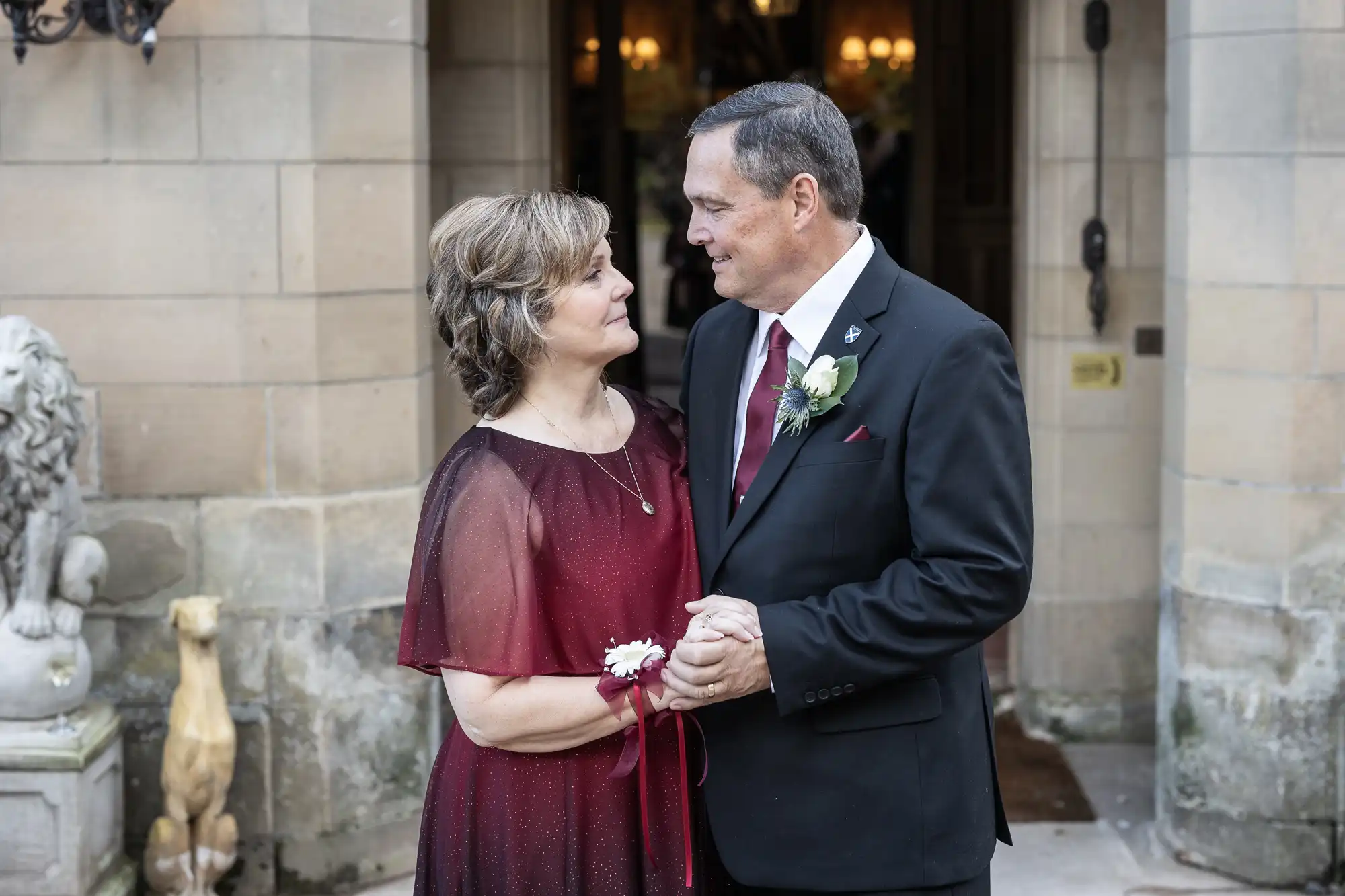 An elderly man and woman stand close, holding hands and looking at each other. They are dressed formally in front of a stone building with decorative statues.