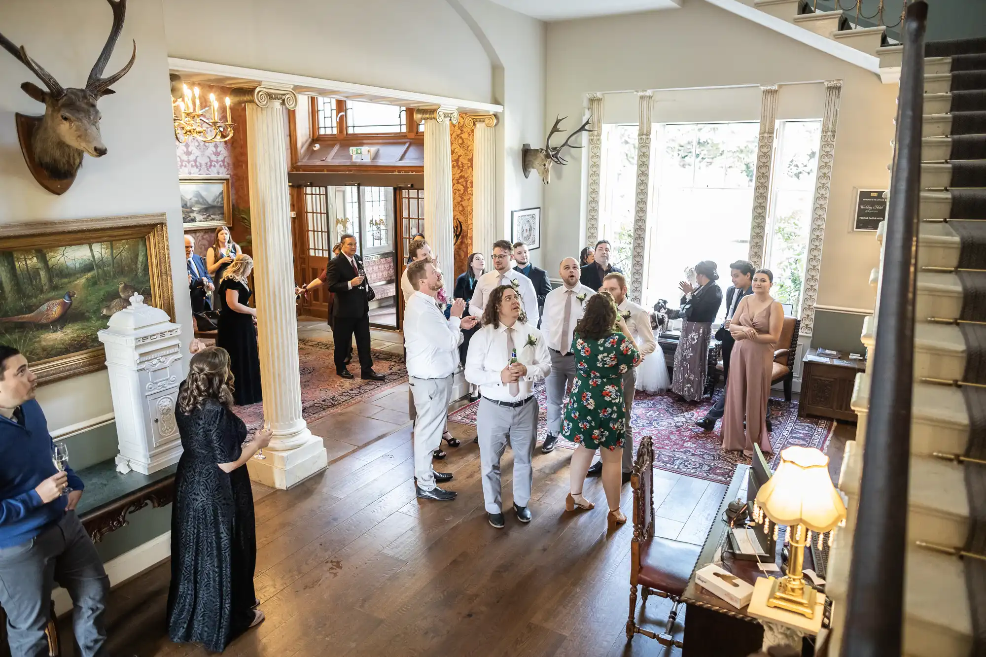 A group of people mingling and chatting in a spacious, elegantly decorated room with large windows, a staircase, and a deer head mounted on the wall.