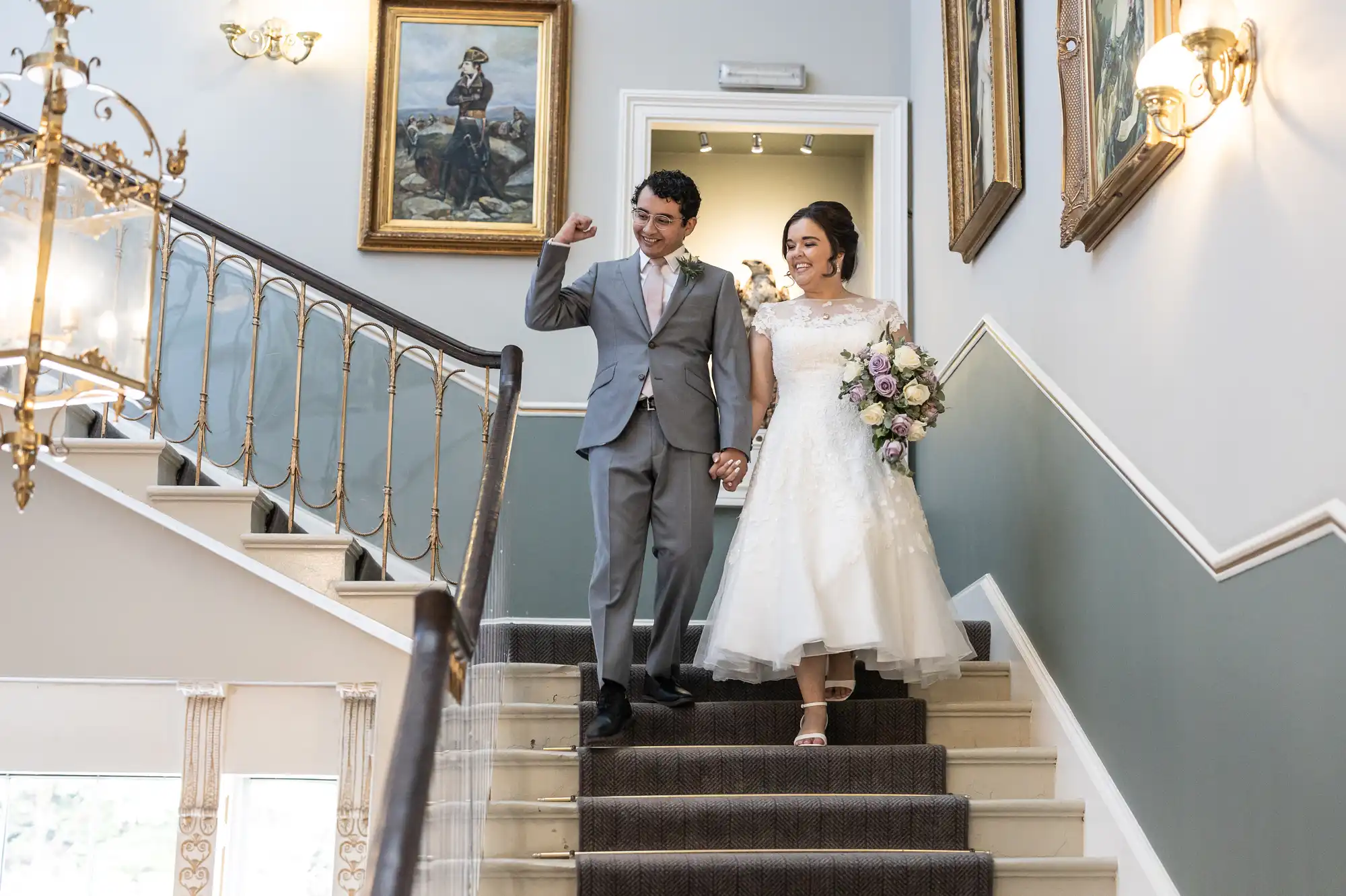 A couple in formal wedding attire descends a staircase, holding hands. The groom wears a gray suit and raises one arm, while the bride wears a white dress and holds a bouquet. Paintings adorn the walls.