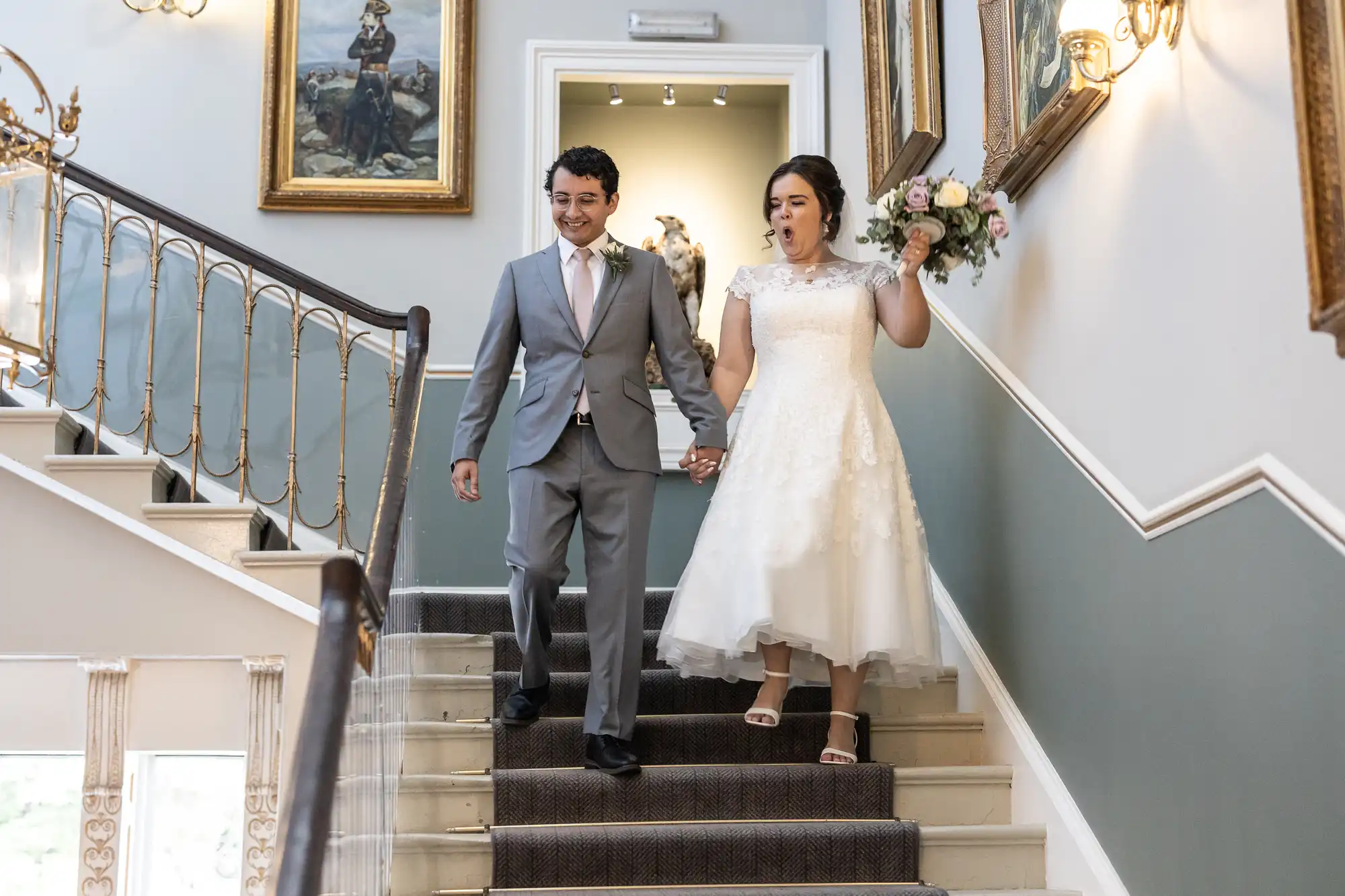 A bride and groom in formal attire hold hands while walking down a staircase. The bride carries a bouquet and they smile at each other. The staircase is adorned with paintings and decorative railings.