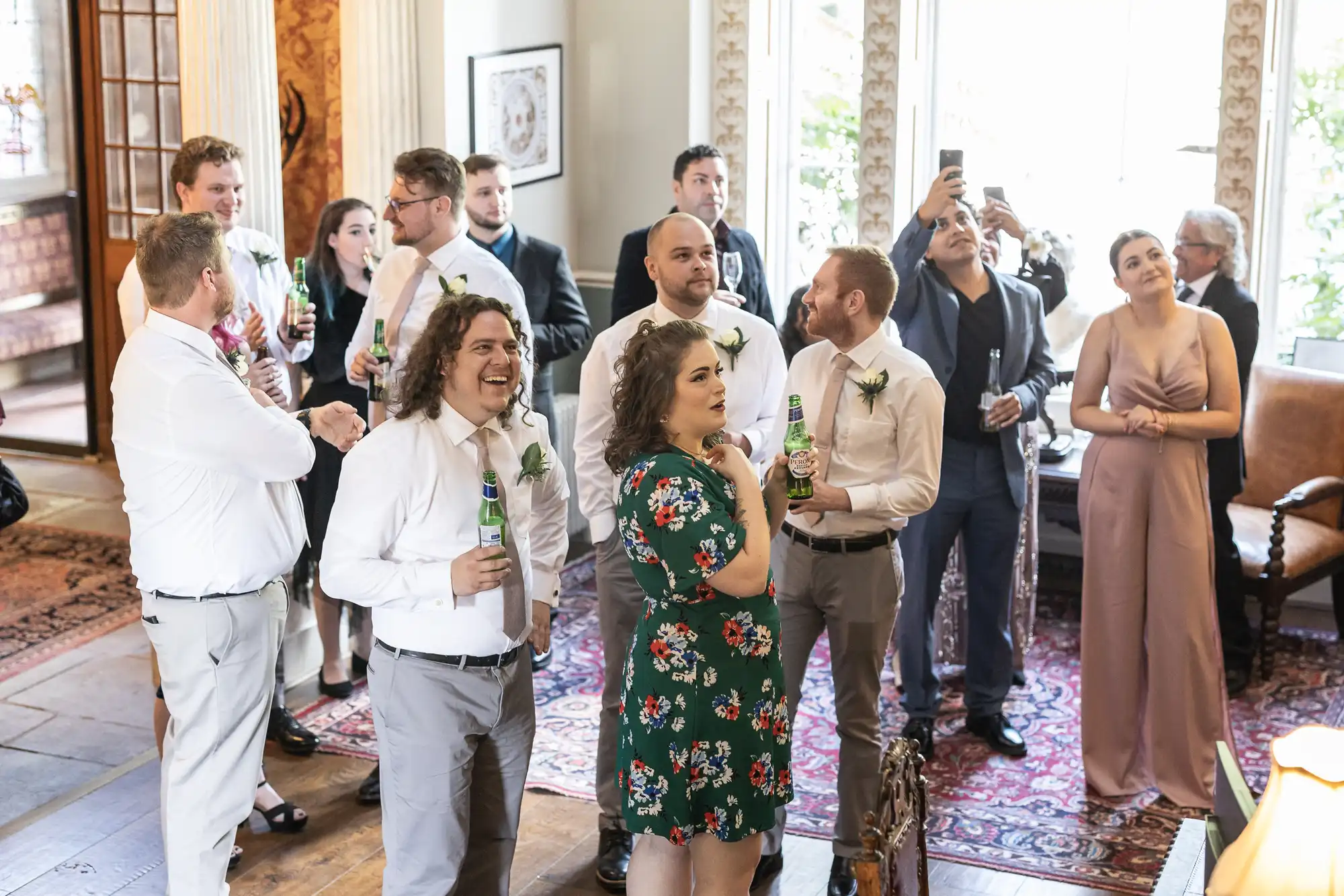 A group of people in semi-formal attire gather indoors with drinks in hand, some looking ahead while others take photos. They are in a well-lit room with ornate decor and large windows.