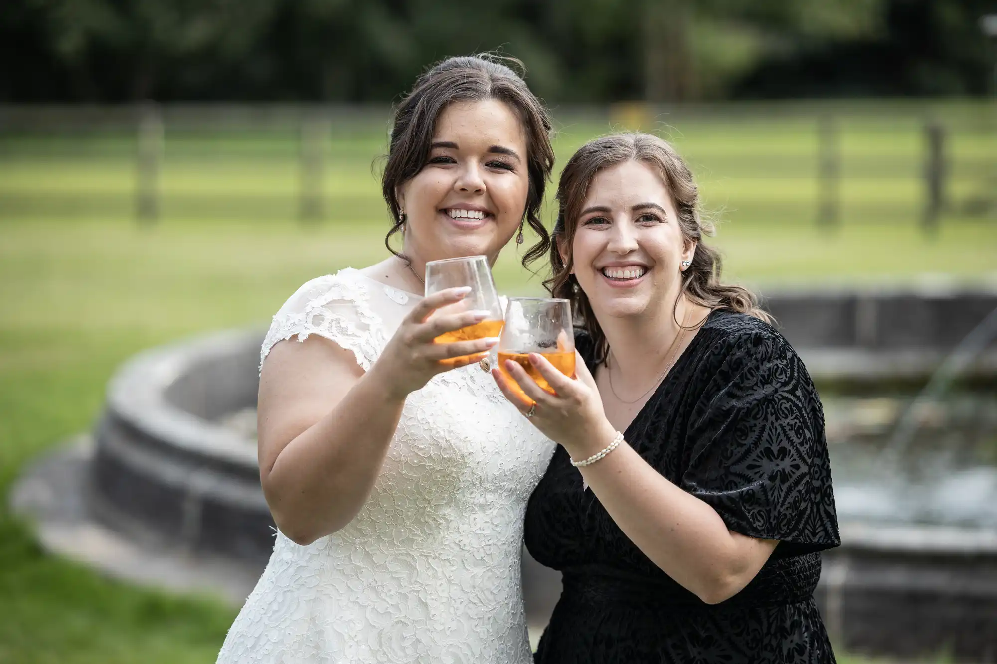 Two women standing outdoors, smiling and holding glasses with orange drinks; one wears a white dress, and the other wears a black dress. There is a grassy area and a stone structure in the background.